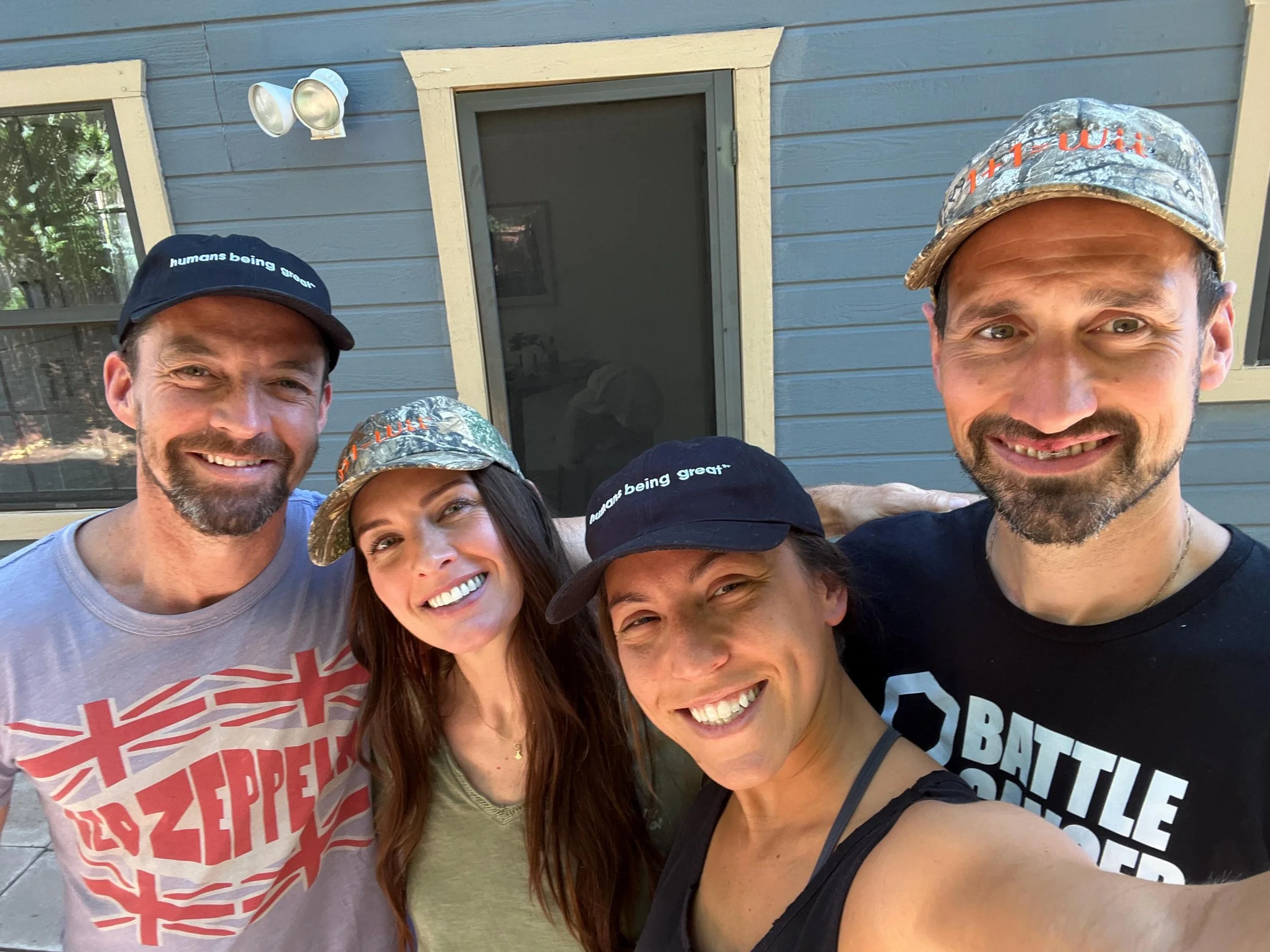 Group of four smiling people taking a selfie outside a house, wearing casual clothes, some with caps and hats, with a blue house in the background.