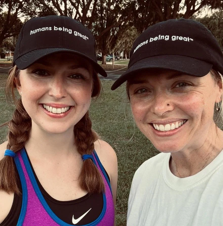Two women smiling outdoors wearing black baseball caps with the phrase 'humans being great™'