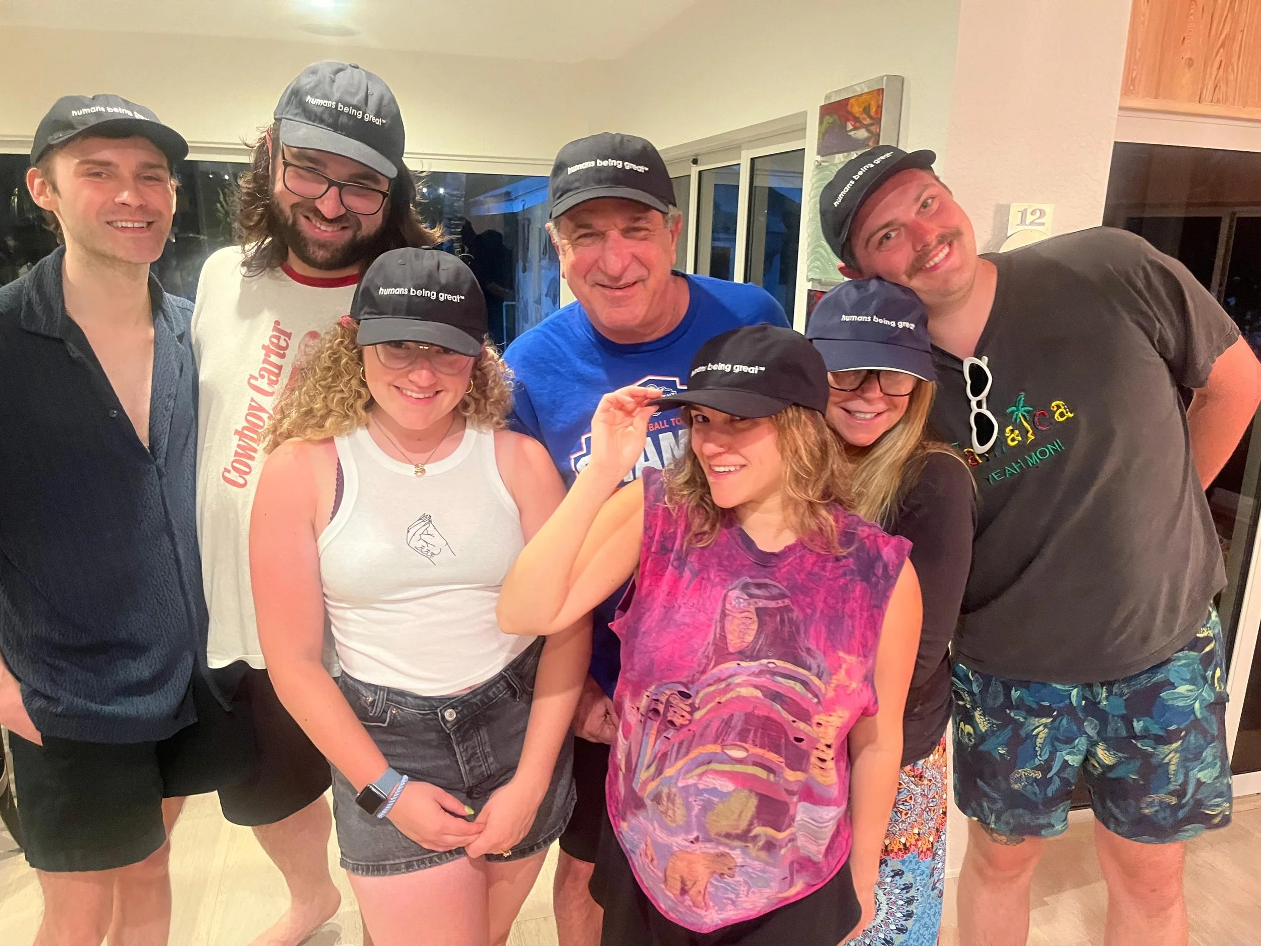 A group of seven people smiling and posing for a photo indoors. They are wearing black caps with the phrase 'humans being great' printed on them. Some are wearing casual summer clothing like t-shirts, tank tops, and shorts.