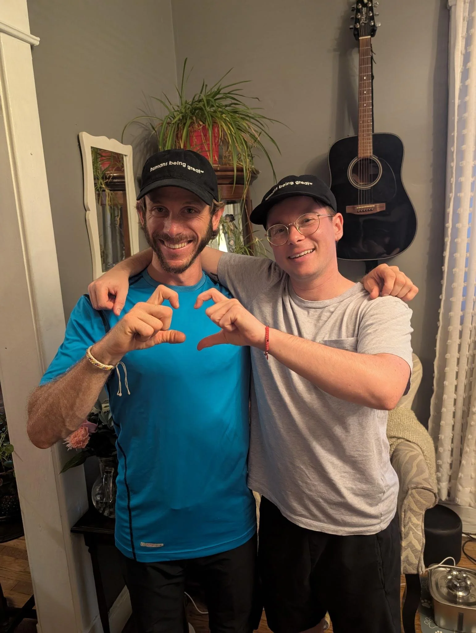 Two smiling men wearing black caps making heart shapes with their hands, standing indoors with a guitar hanging on the wall behind them.