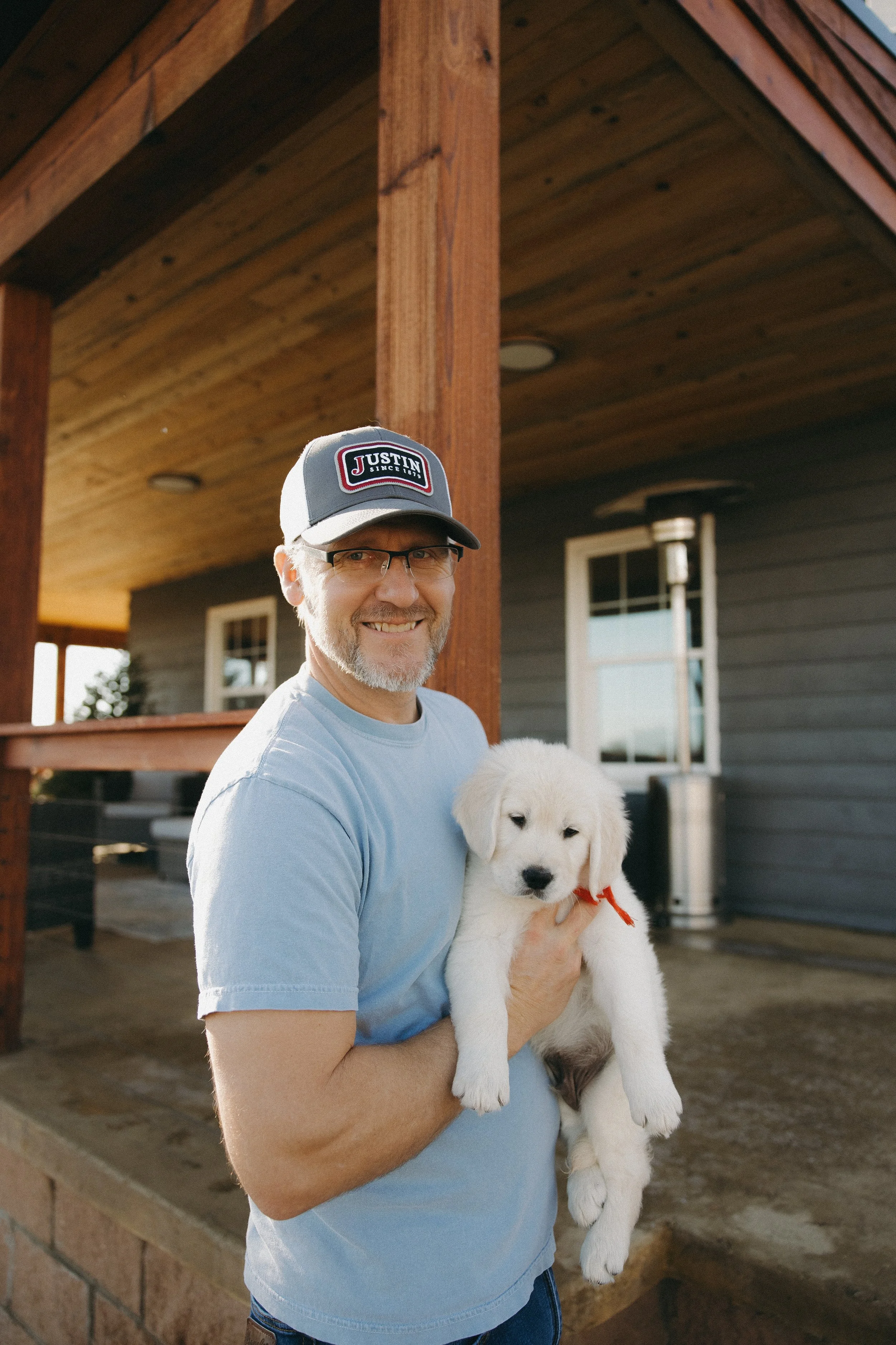 A man with gray hair and glasses is sitting in a chair holding a happy golden retriever puppy near a red wall.