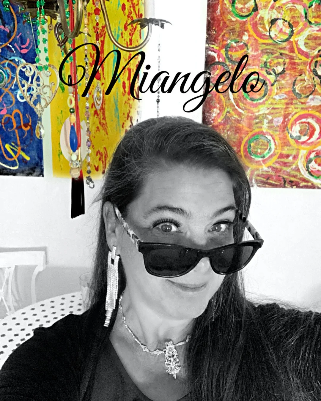 A woman with long dark hair, wearing sunglasses, earrings, and a necklace, taking a selfie. Behind her are colorful abstract paintings and jewelry displays. The text 'Miangelo' is across the top.
