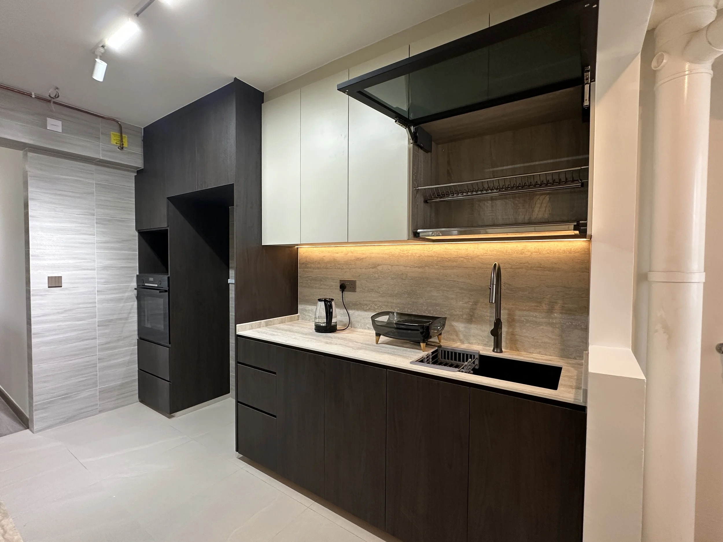Modern kitchen with dark lower cabinets, white upper cabinets, beige backsplash, and built-in appliances. There is a black kettle on the counter, a small dish rack, and a wall-mounted pull-up bar.