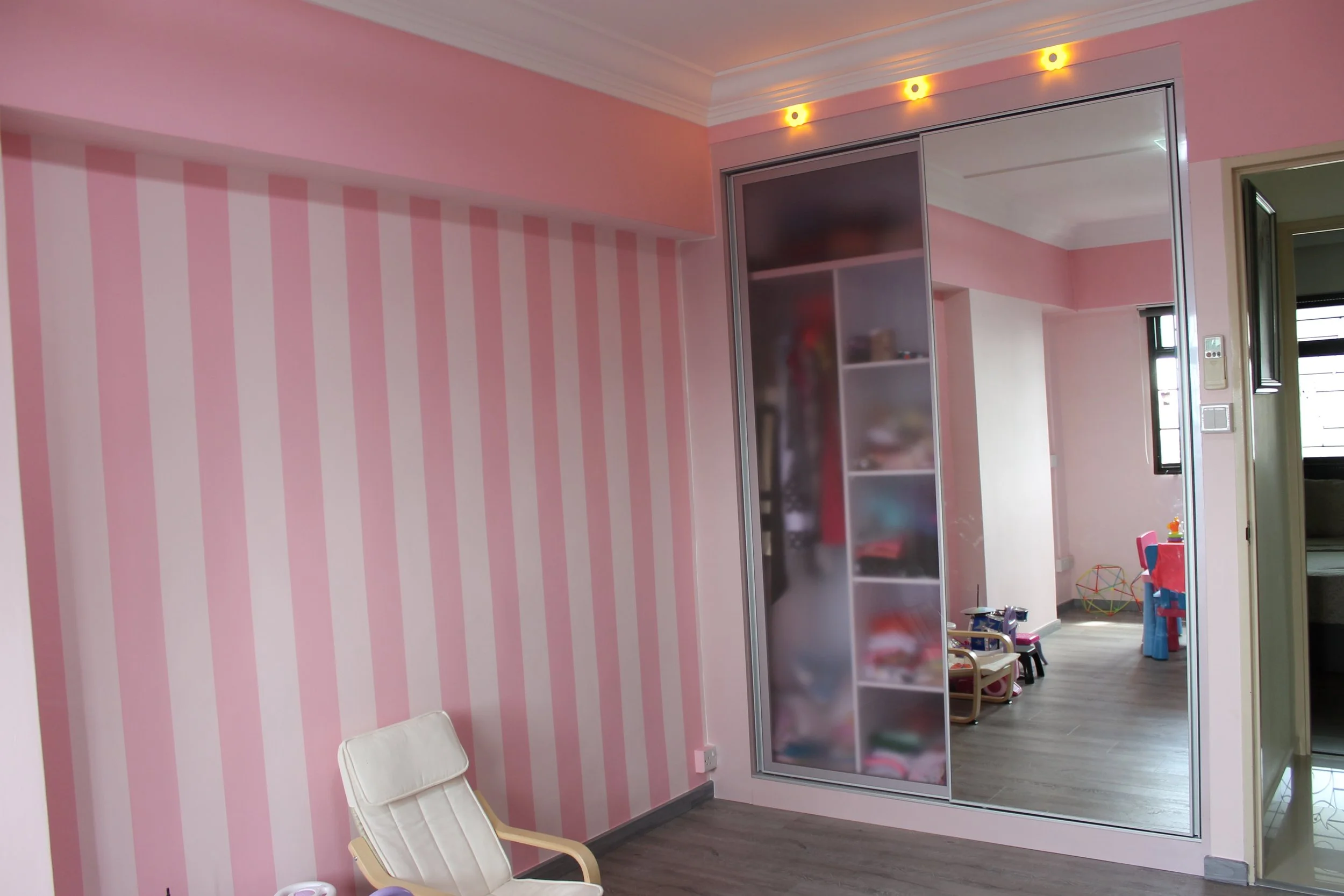 Interior of a room with pink and white striped wall, a white chair, and a sliding glass door reflecting a playroom with toys and children's furniture.