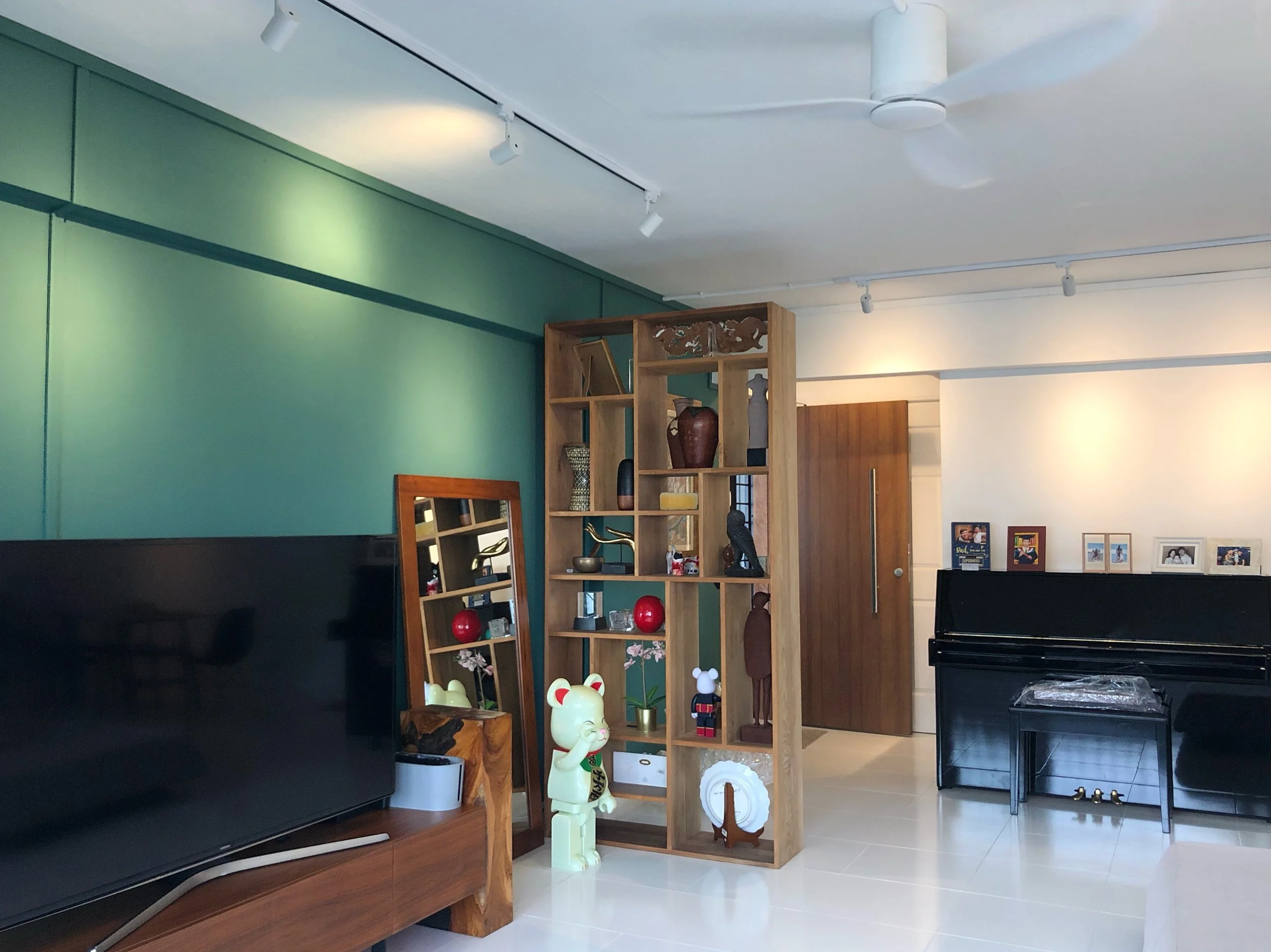 Living room with a black TV, wooden shelves filled with decorative items, a mirror, a few framed photos, and a black piano against a white wall. Green accent wall and ceiling fan visible.