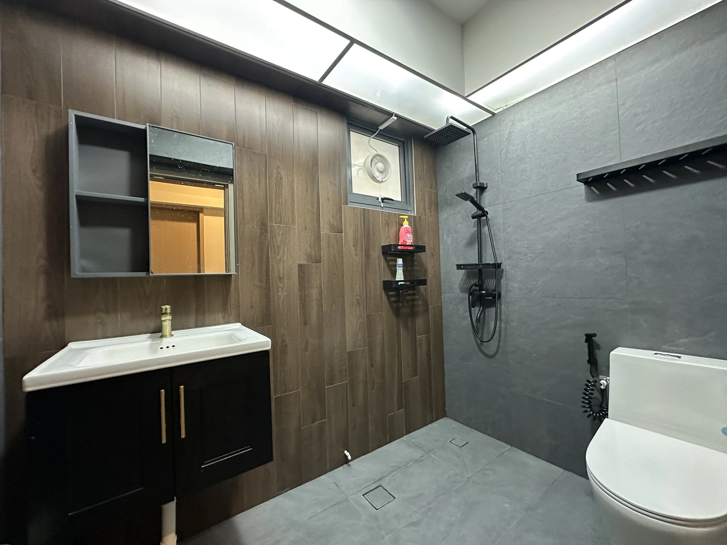 Bathroom with wood-effect and gray tiles, wall-mounted mirror, black vanity with gold handles, white vessel sink, showerhead, curtain rod, small window with exhaust fan, and a white toilet.