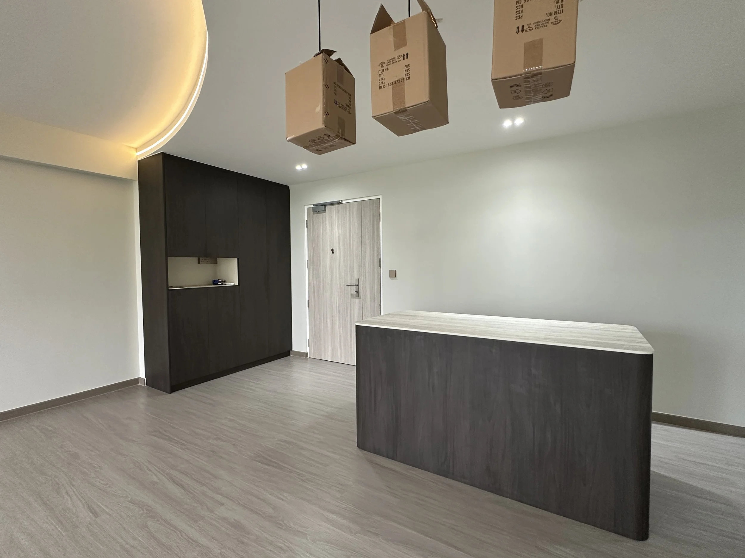 Interior of a modern room with light-colored wooden flooring, white walls, and a dark wooden cabinet. Three cardboard boxes are hanging from the ceiling.