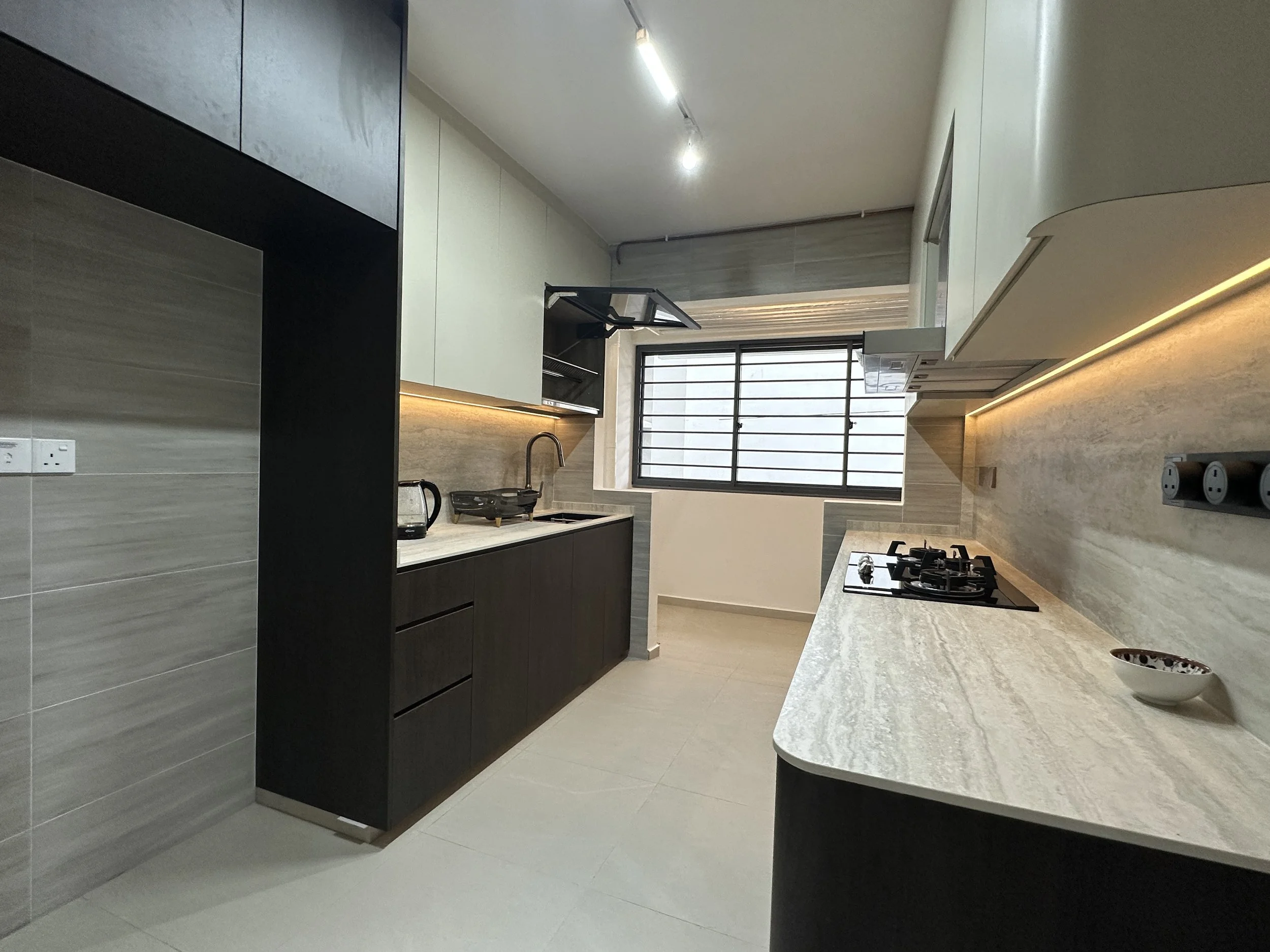 A modern kitchen with white countertops, black cabinets, and a large window with horizontal bars. There are some kitchen appliances and utensils on the counter, and the room is well-lit with overhead lighting.