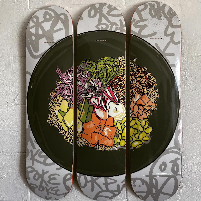 Three skateboard decks displaying a vibrant illustration of a poke bowl, featuring various vegetables and toppings, against a white wall with graffiti-style text in the background.