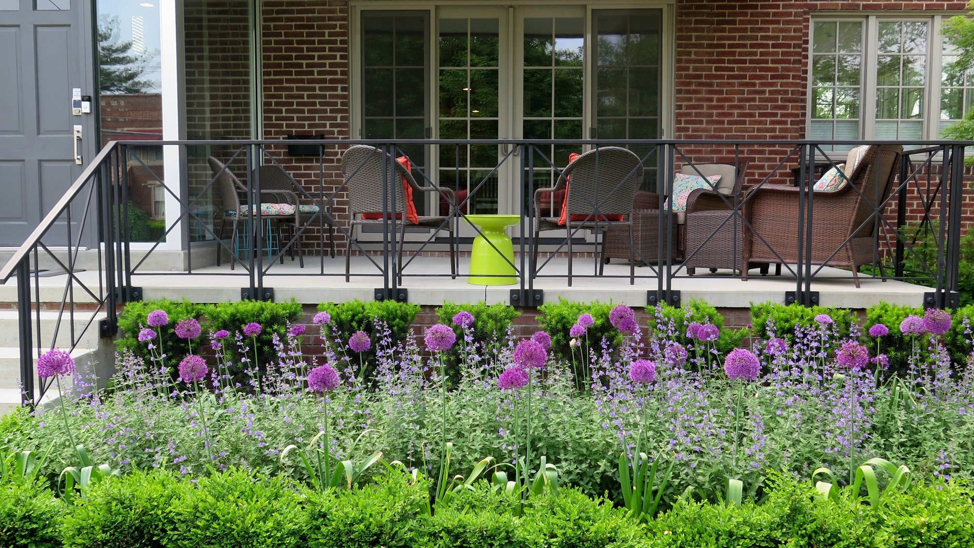 The modern concrete floor with stylish modern black railing is decorated in front with a layered row of plants including a row of each. low boxwood hedge, soft light purple nepeta, with a sprinkling of brighter purple allium balls, and a bright green