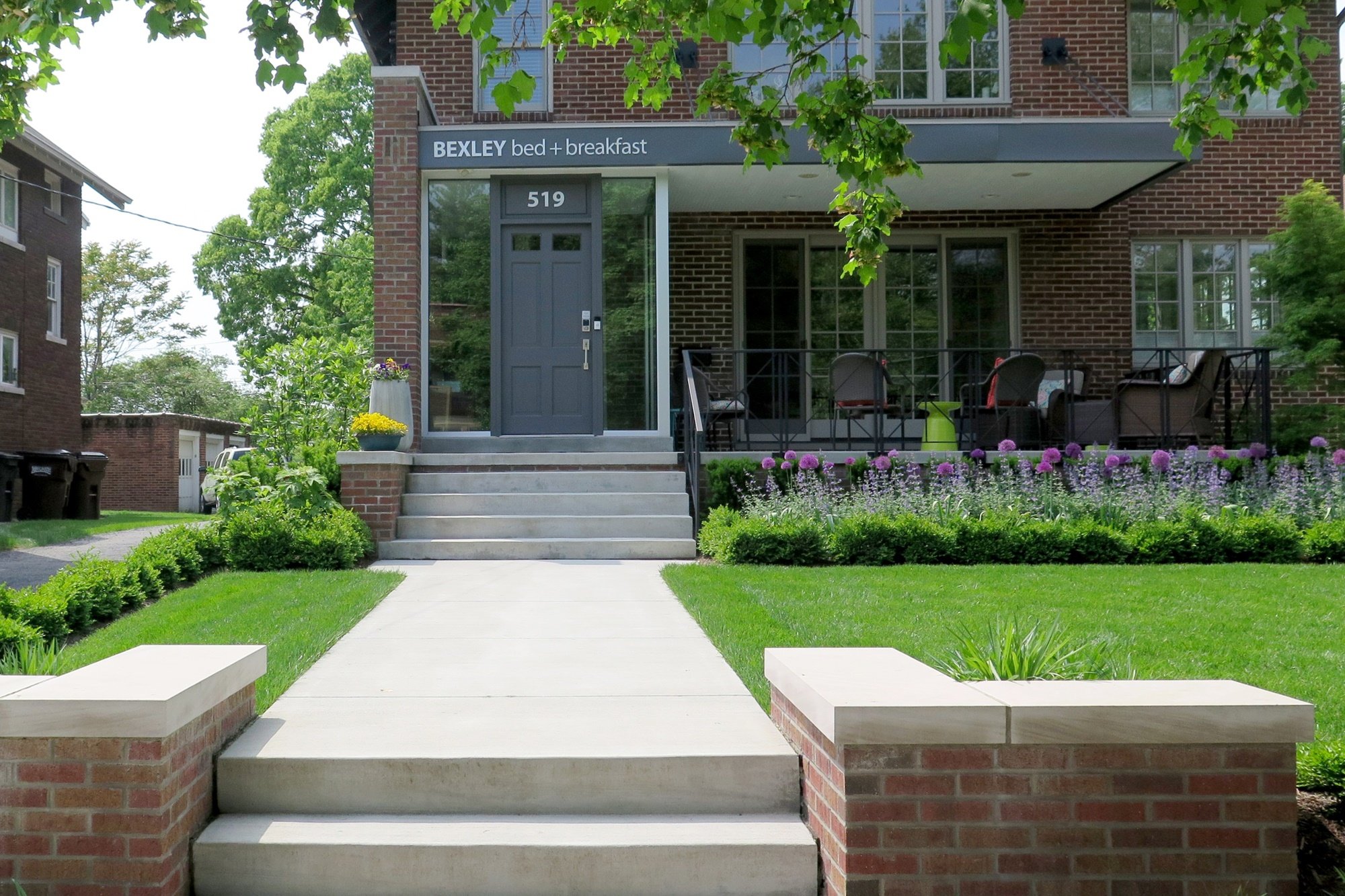Two brick pillars sit on either side of a wide modern light wash concrete pathway to the steps of the front door of the contemporary remodeled brick building