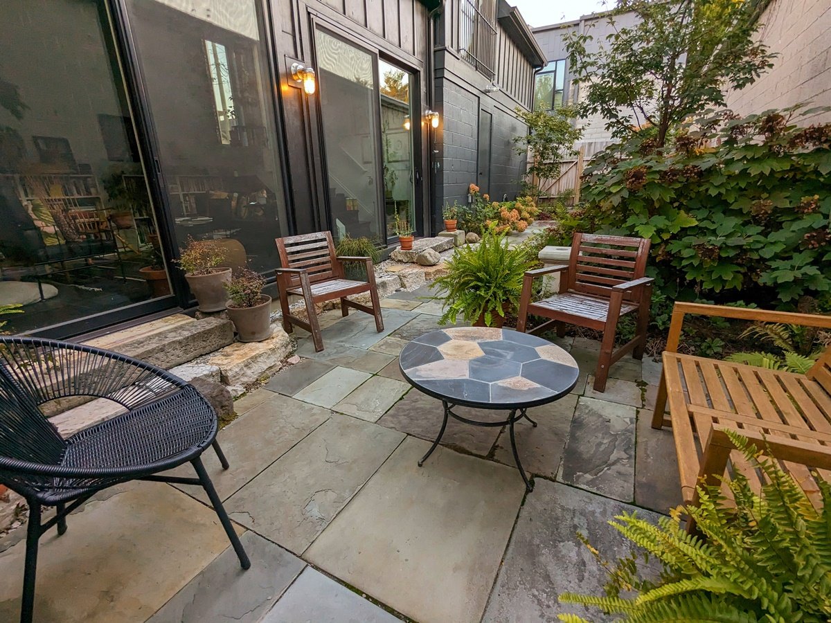 A small sitting area with a wood-backed bench and chair to the right and a contemporary black wire chair to the left of it sit nicely on this cozy bluestone patio made with odd-sized pieces