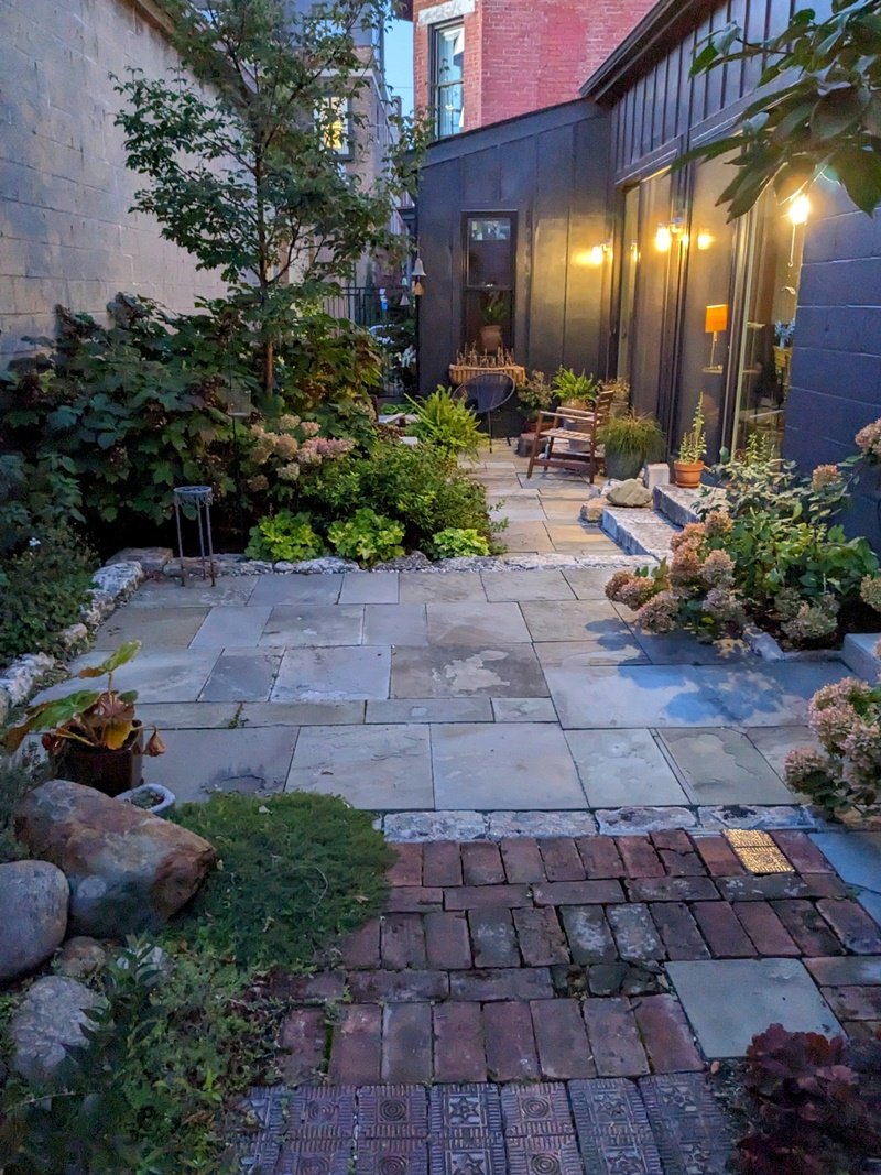 A long, narrow patio made up of many types of materials, such as bluestone, brick, and limestones, is sandwiched between two buildings with soft, varied plantings on the sides