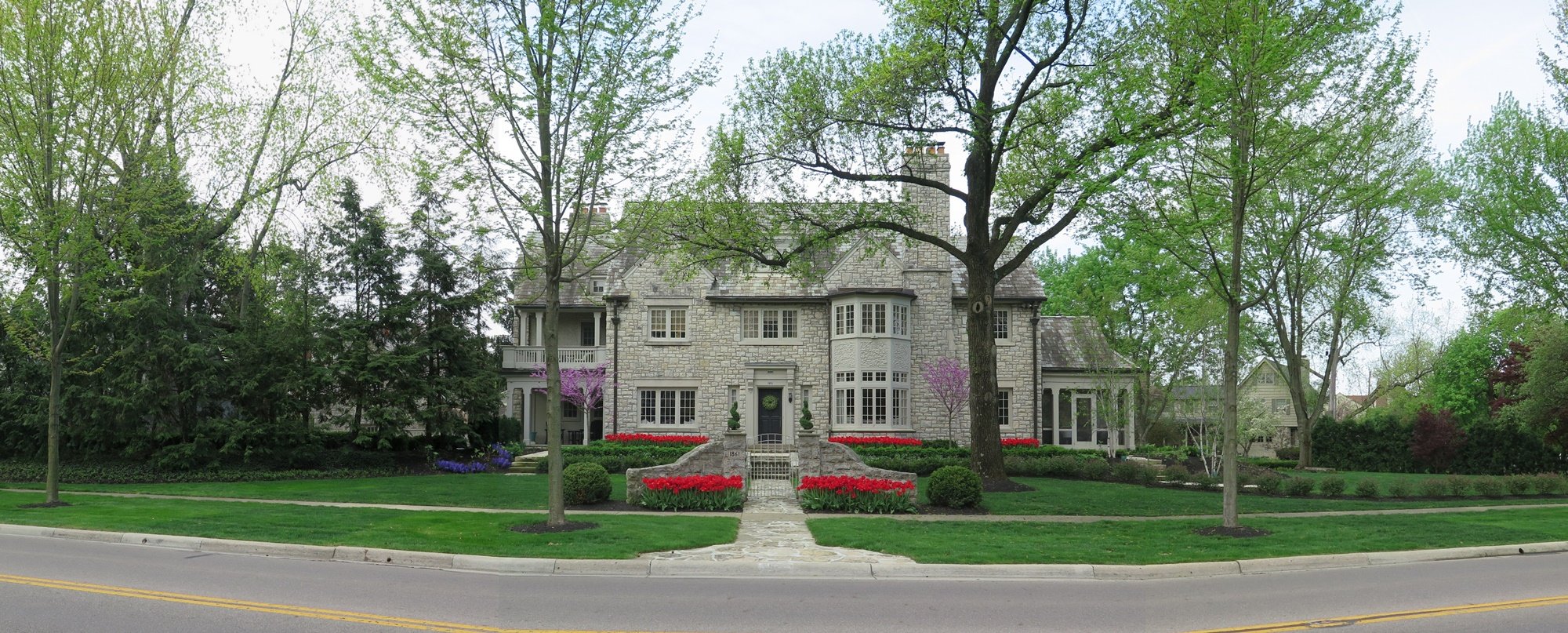 Street view of two-story limestone house with lush lawn and large trees and bright spots of red tulips in front of the house