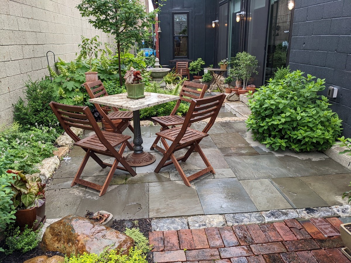 A small square café table and four wood folding chairs sit on a bluestone square made of various sizes of stone and extending beyond with various types and sizes of brick and other materials
