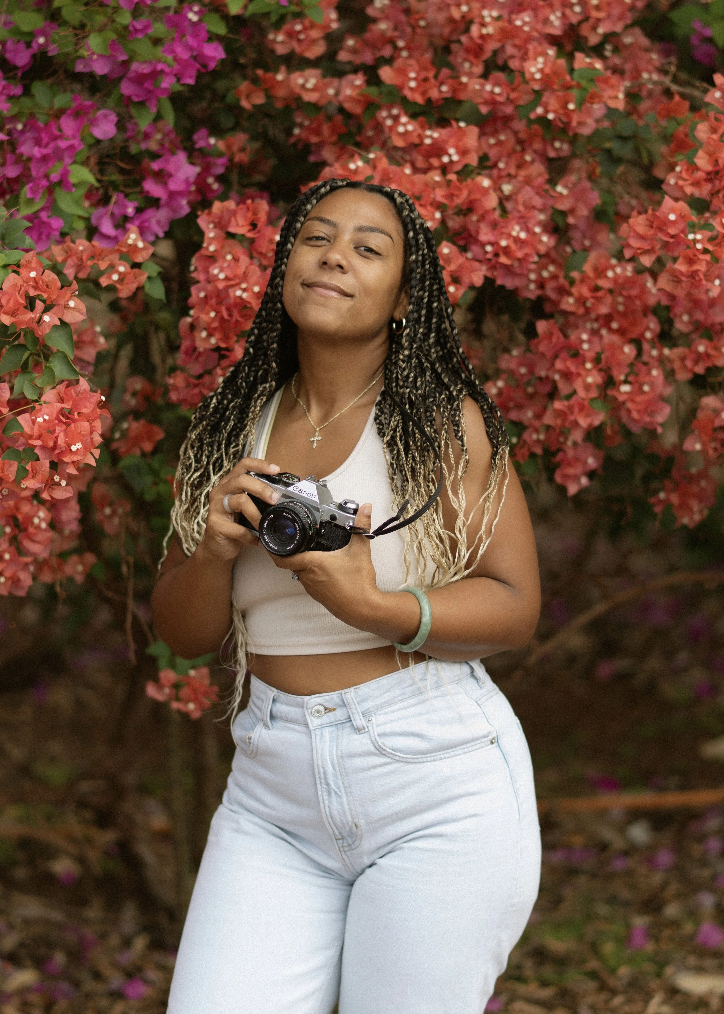 A woman holding a camera standing in front of pink and orange bougainvillaea flowers.