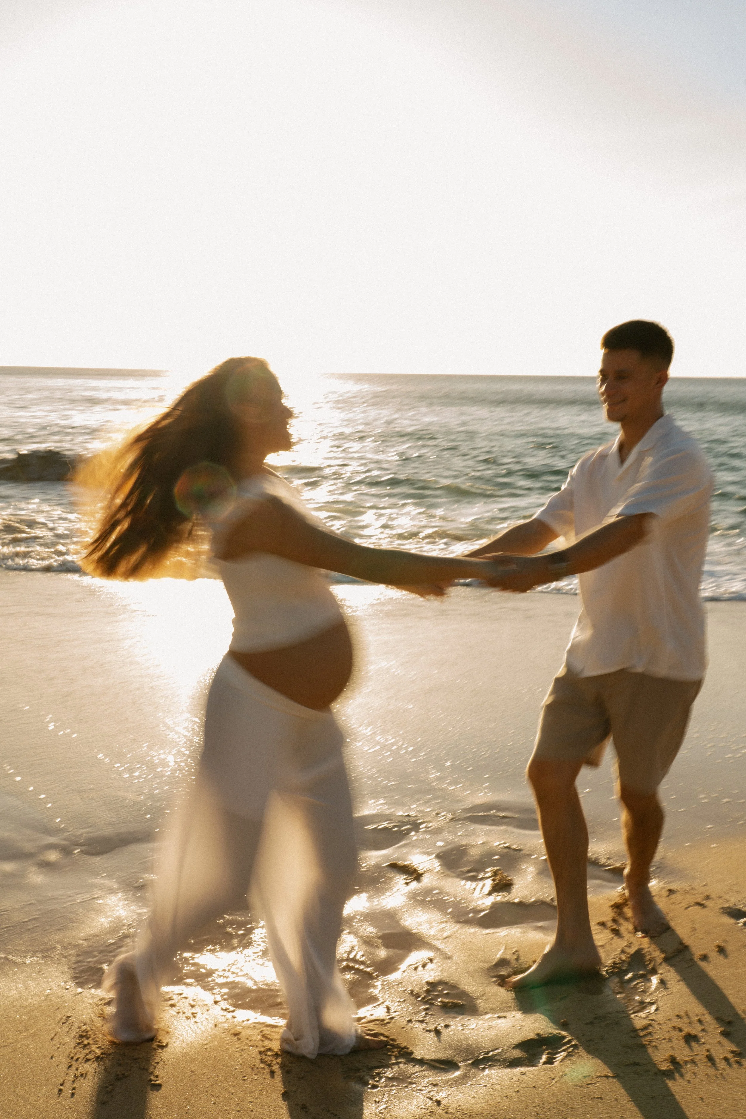 A couple holding hands and dancing on the beach during sunset, with the ocean in the background.