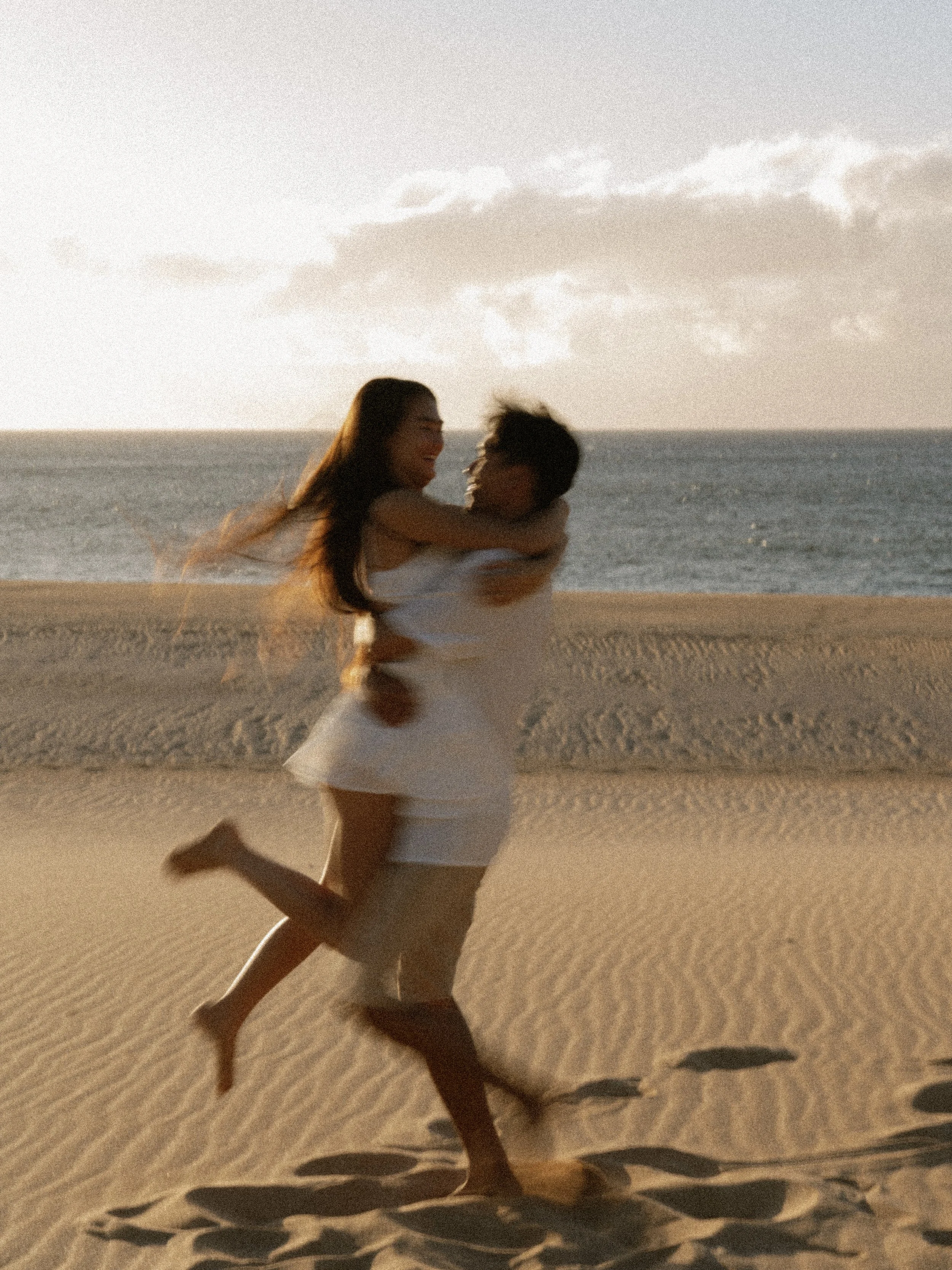 A couple on a sandy beach during sunset, with the woman being lifted by the man. They are both smiling, and the ocean and sky are visible in the background.