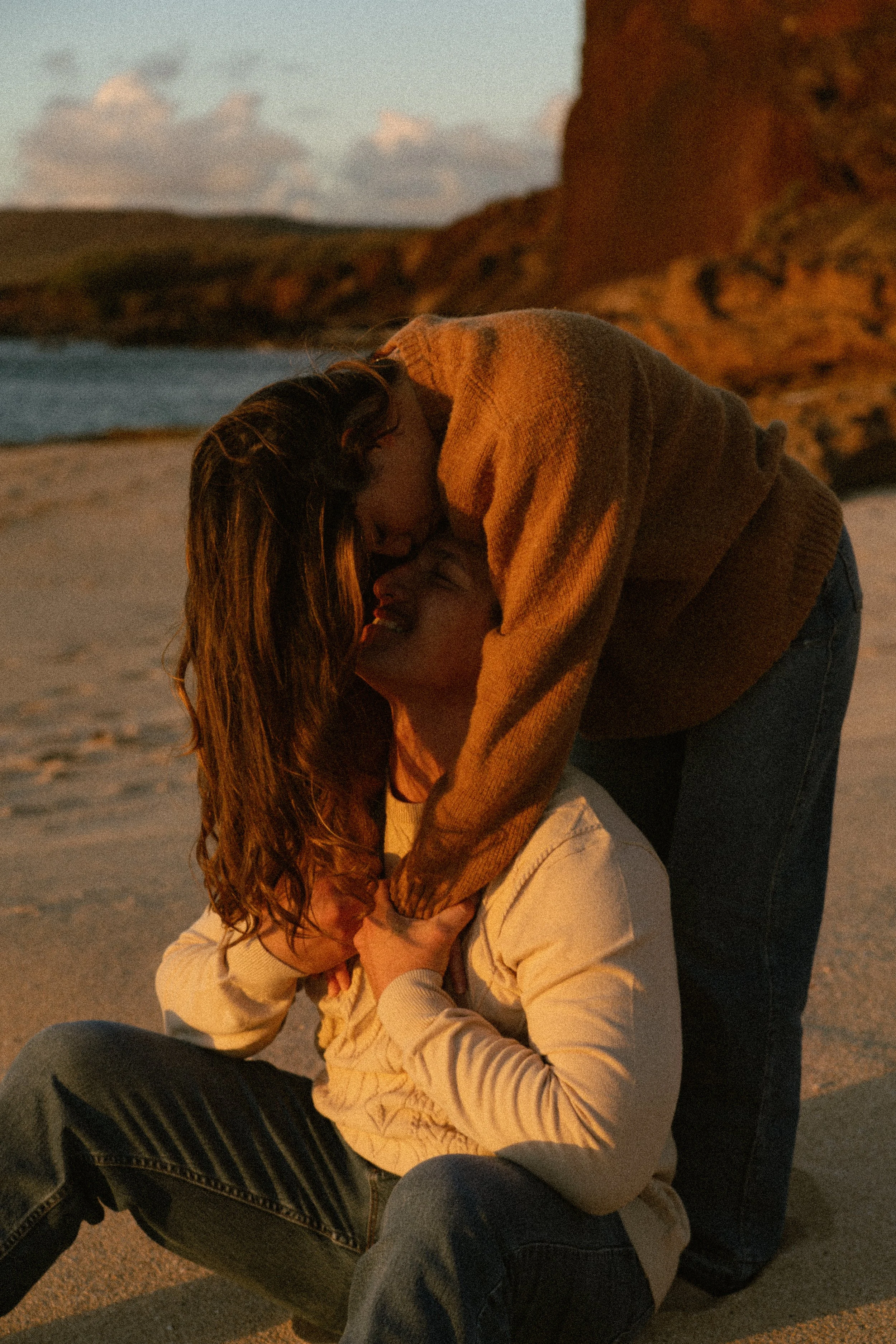 A couple on a beach during sunset, with the woman sitting on the sand and the man leaning over, touching her face as they share a tender moment.