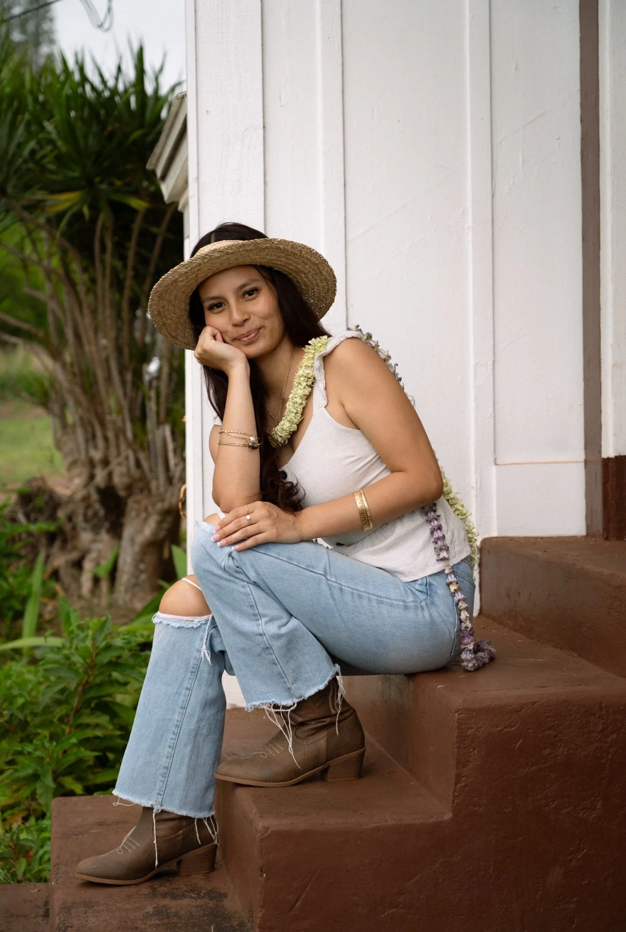 A woman sitting on outdoor stairs, wearing a straw hat, light-colored sleeveless top, ripped jeans, and brown boots, with jewelry and flower garland accessories.