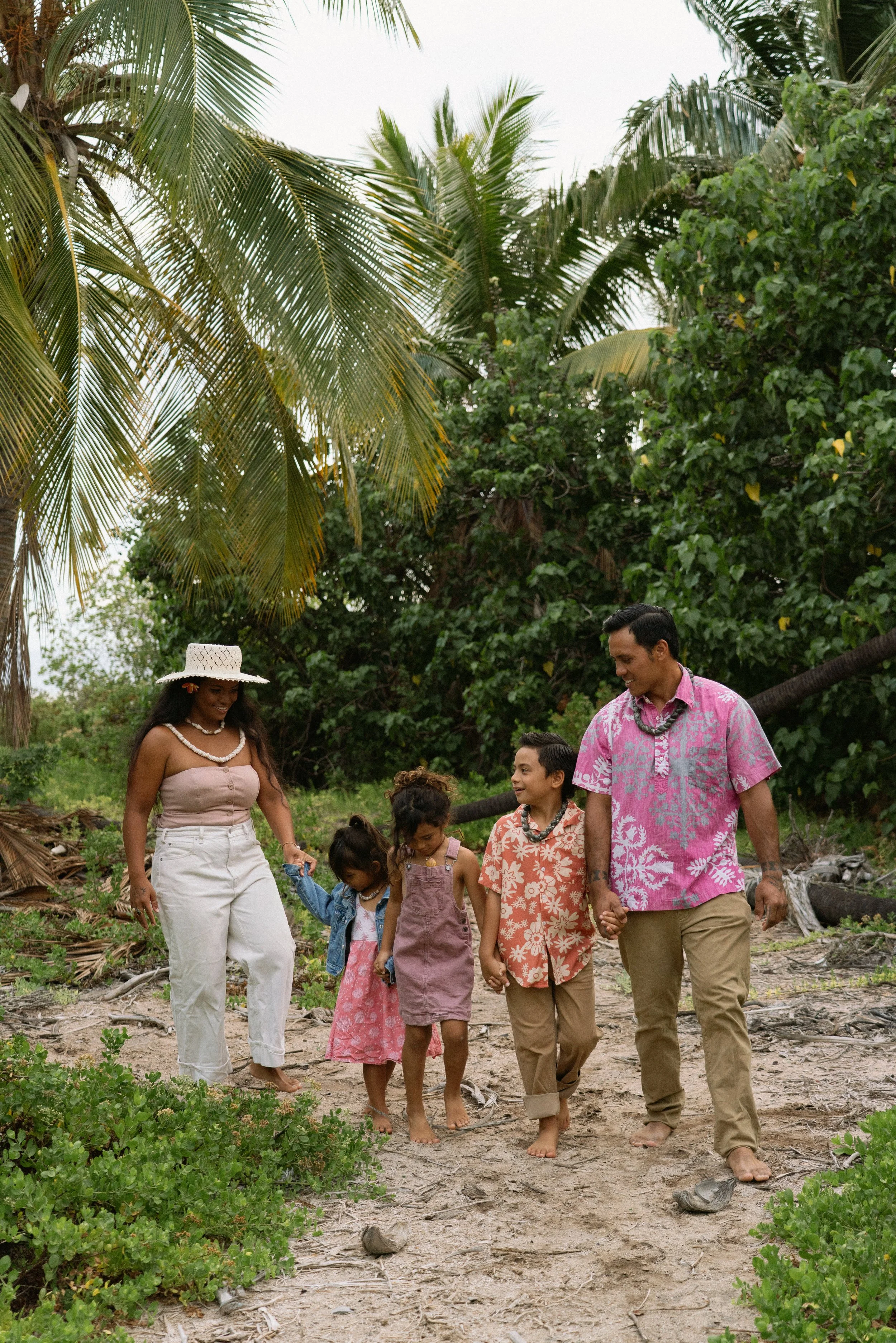 A family walking barefoot on a sandy trail through a tropical setting with palm trees and lush green foliage, holding hands and smiling.