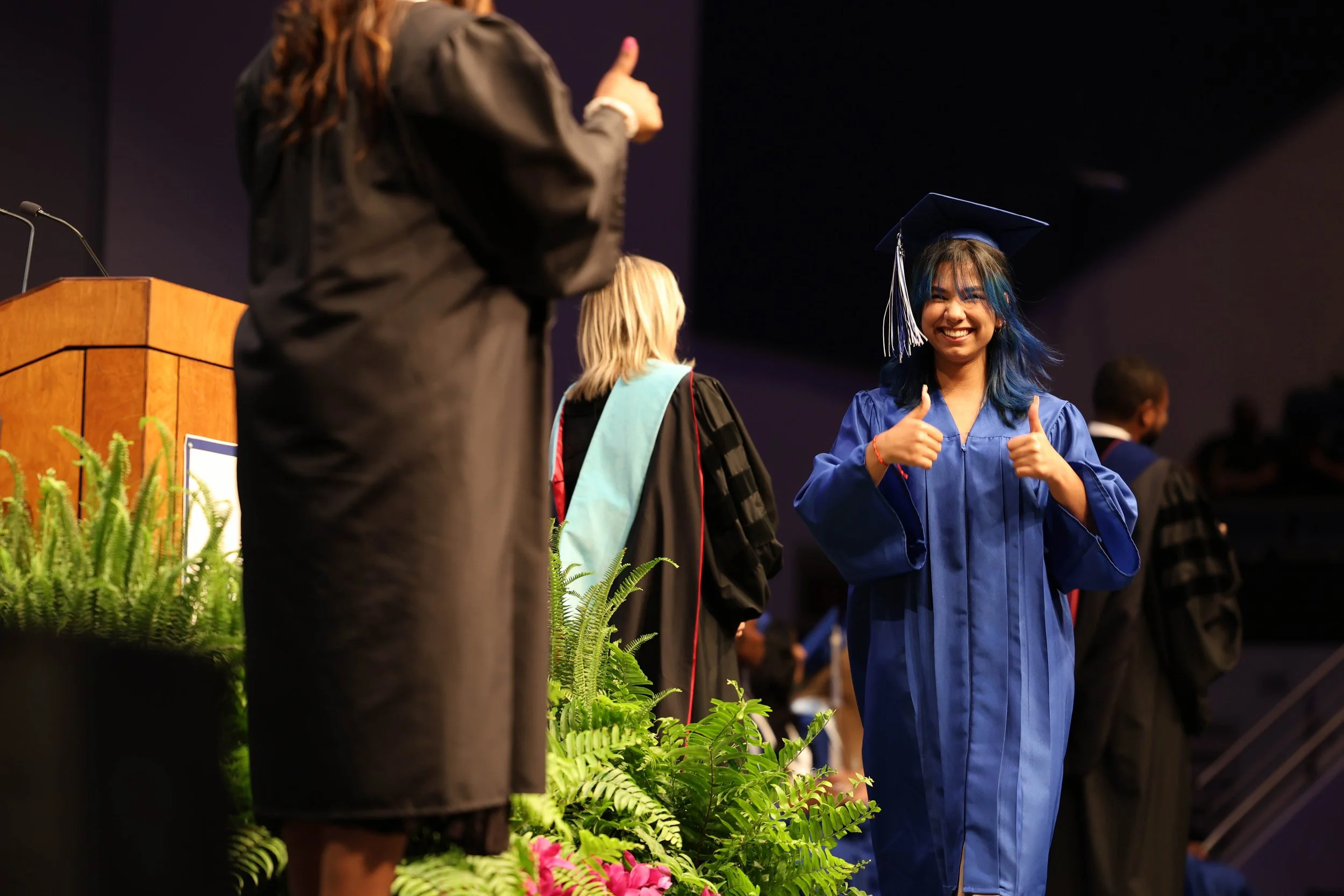 Young woman with blue hair in a graduation cap and gown smiling and giving a thumbs-up at a graduation ceremony.