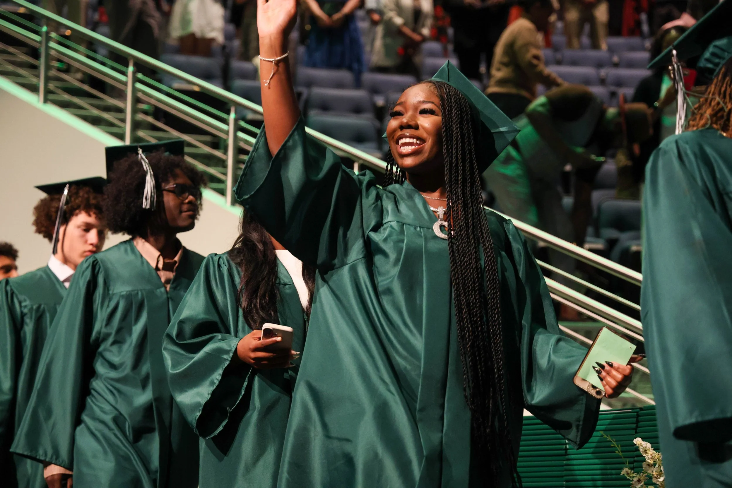 A woman in a green graduation cap and gown smiling and waving at a graduation ceremony, surrounded by other graduates in similar attire, some holding smartphones.