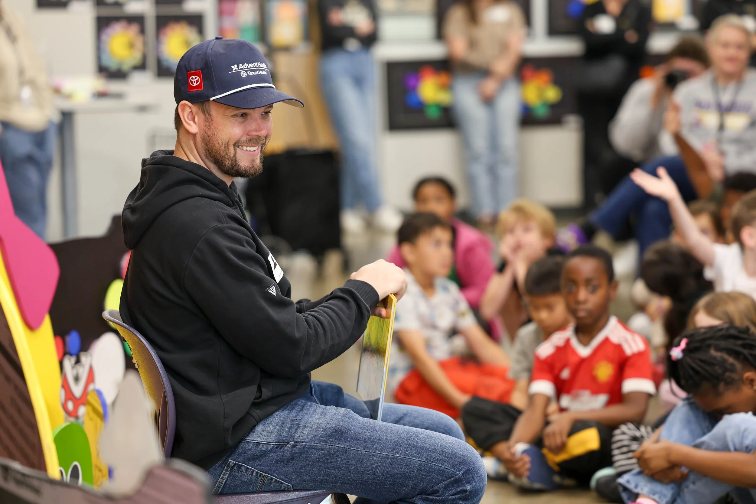A man with a beard and wearing a navy blue cap and black hoodie sitting in front of children in a classroom or auditorium listening attentively.