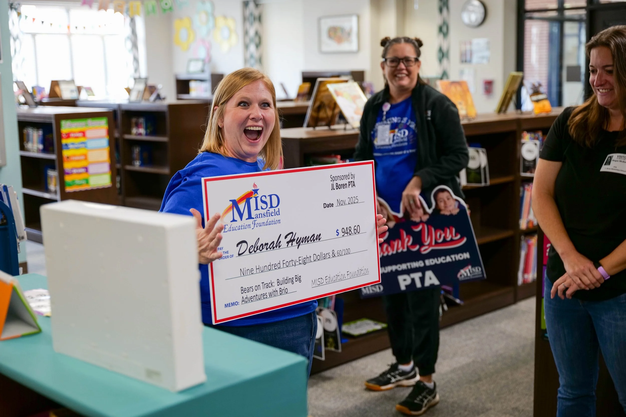 Woman holding large donation check while smiling in a bookstore with other women around, colorful decorations and bookshelves in background.