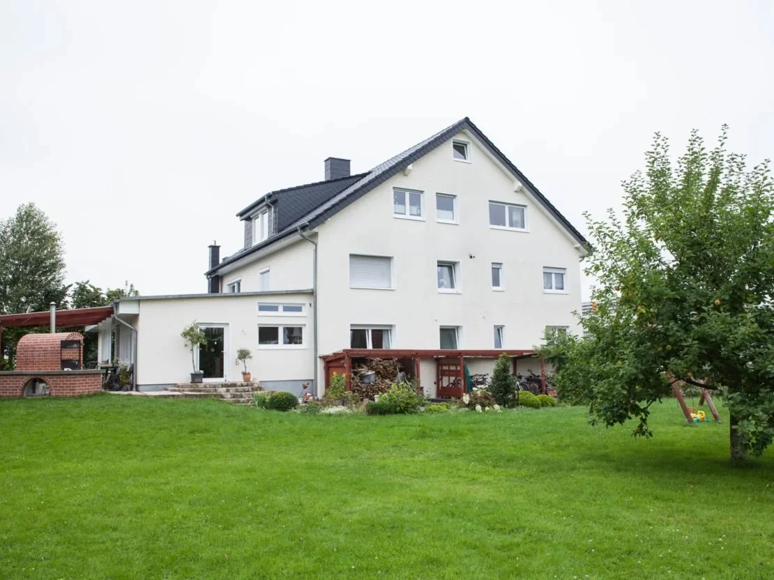 Großes, weißes Einfamilienhaus mit Satteldach, Garten mit grünem Rasen, Baum und überdachtem Backofen im Hintergrund.