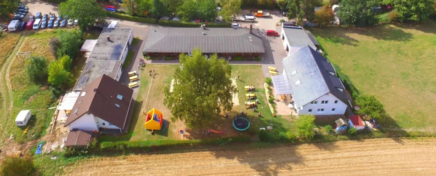 Luftaufnahme eines Gebäudekomplexes mit mehreren Häusern, umgeben von einer Wiese und einem Parkplatz mit Autos. Im Gartenbereich sind ein Baum, ein Trampolin und ein Hüpfburg zu sehen.