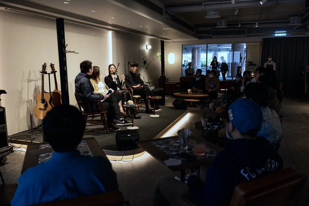Four people are sitting on stage in a panel discussion at a cafe or lounge, with guitars leaning against the wall behind them. Audience members are seated in front, listening, with some wearing face masks. The room has dim lighting and a cozy atmosphere.