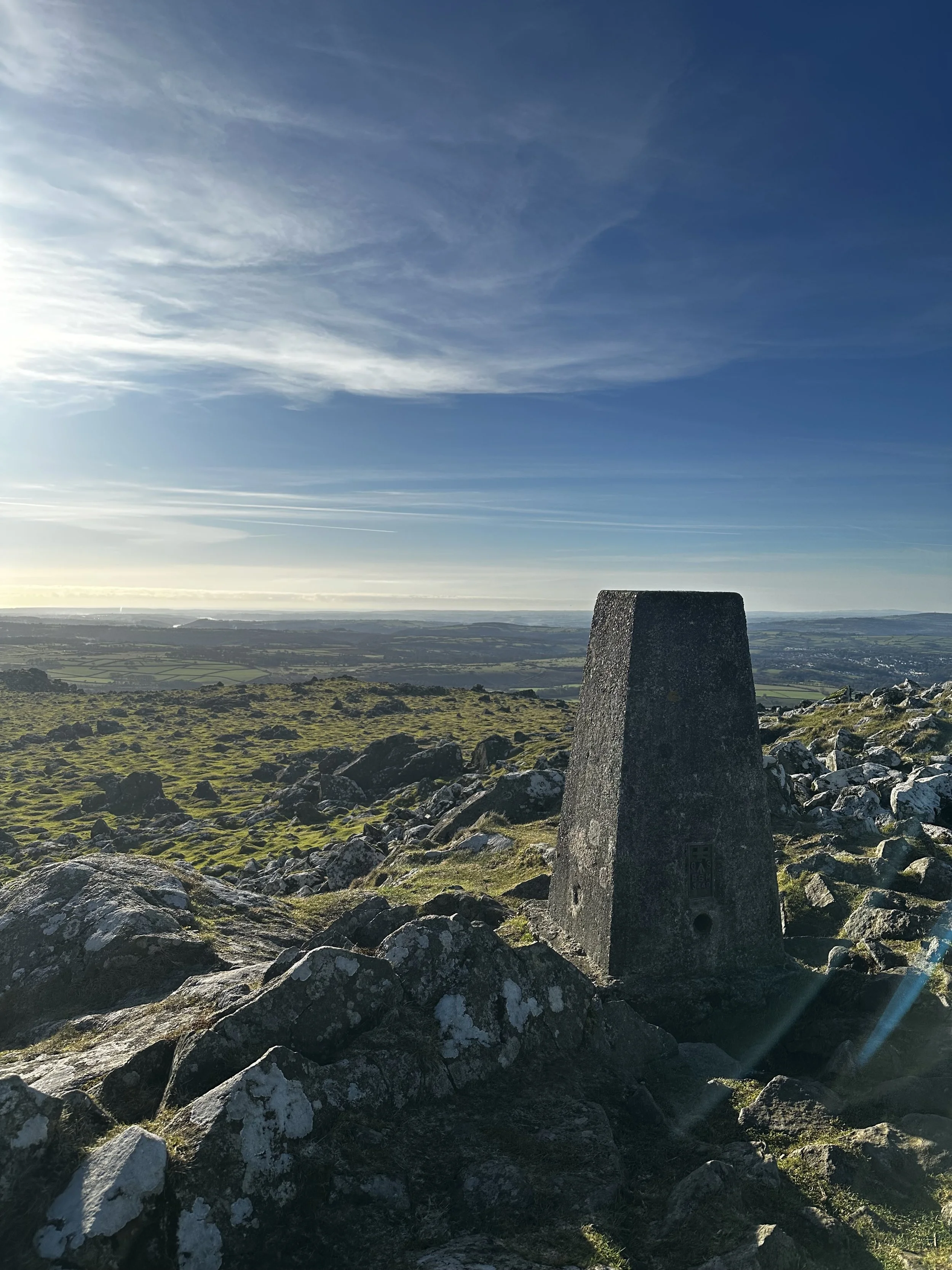 A view of a trig point on the top of Cox Tor, looking over the Tamar river in the distance