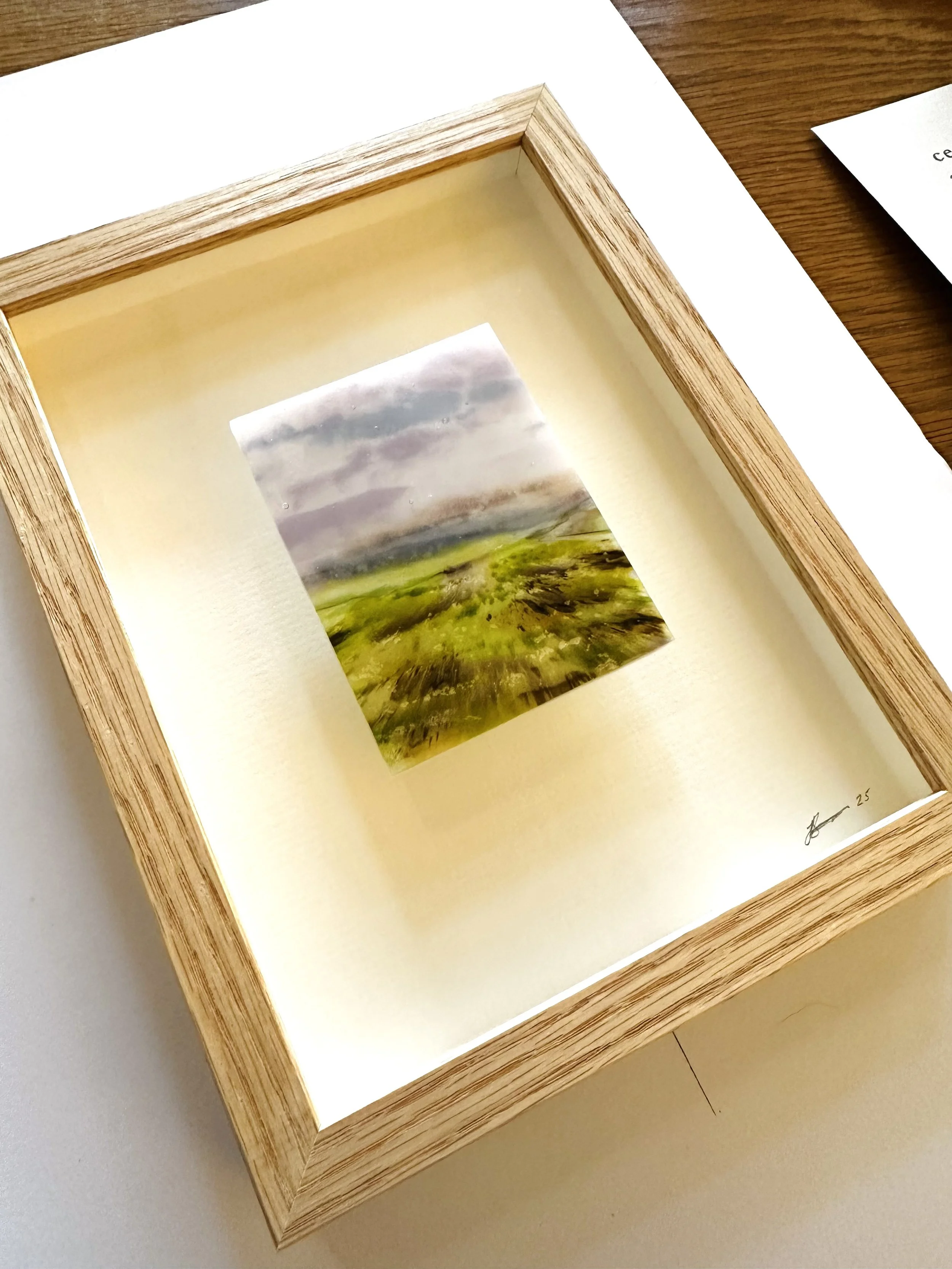 A close up of an oak picture frame flat on a table, with a glass landscape mounted onto the frame glass.