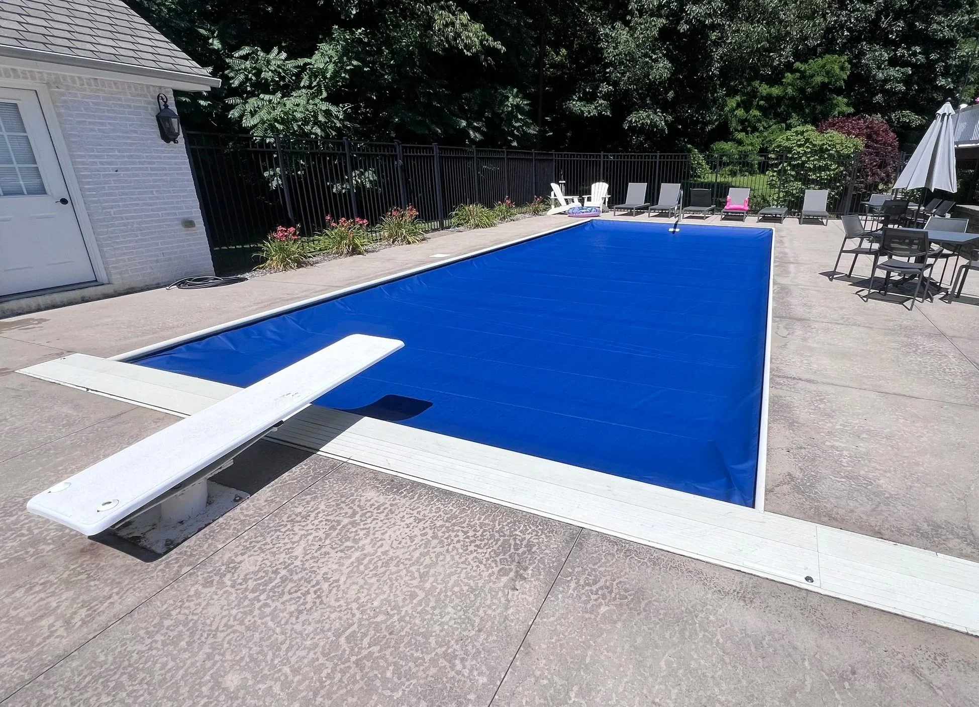 Empty swimming pool with a blue cover in a backyard, surrounded by lounge chairs, patio furniture, and lush greenery.