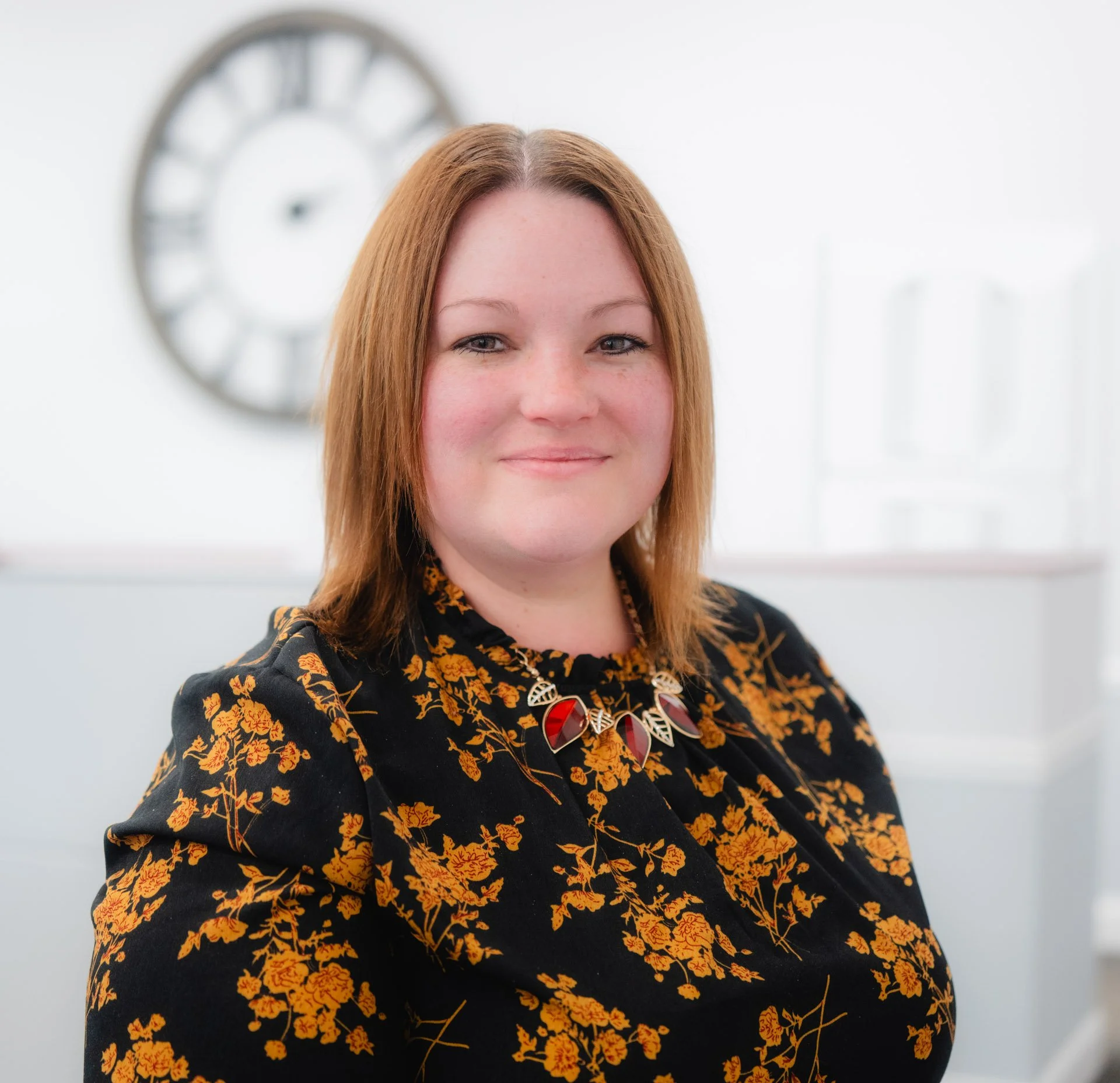 A woman with shoulder-length reddish hair smiling in a bright office space. She is wearing a black dress with a yellow floral pattern and a colorful necklace. A large clock is visible in the background.
