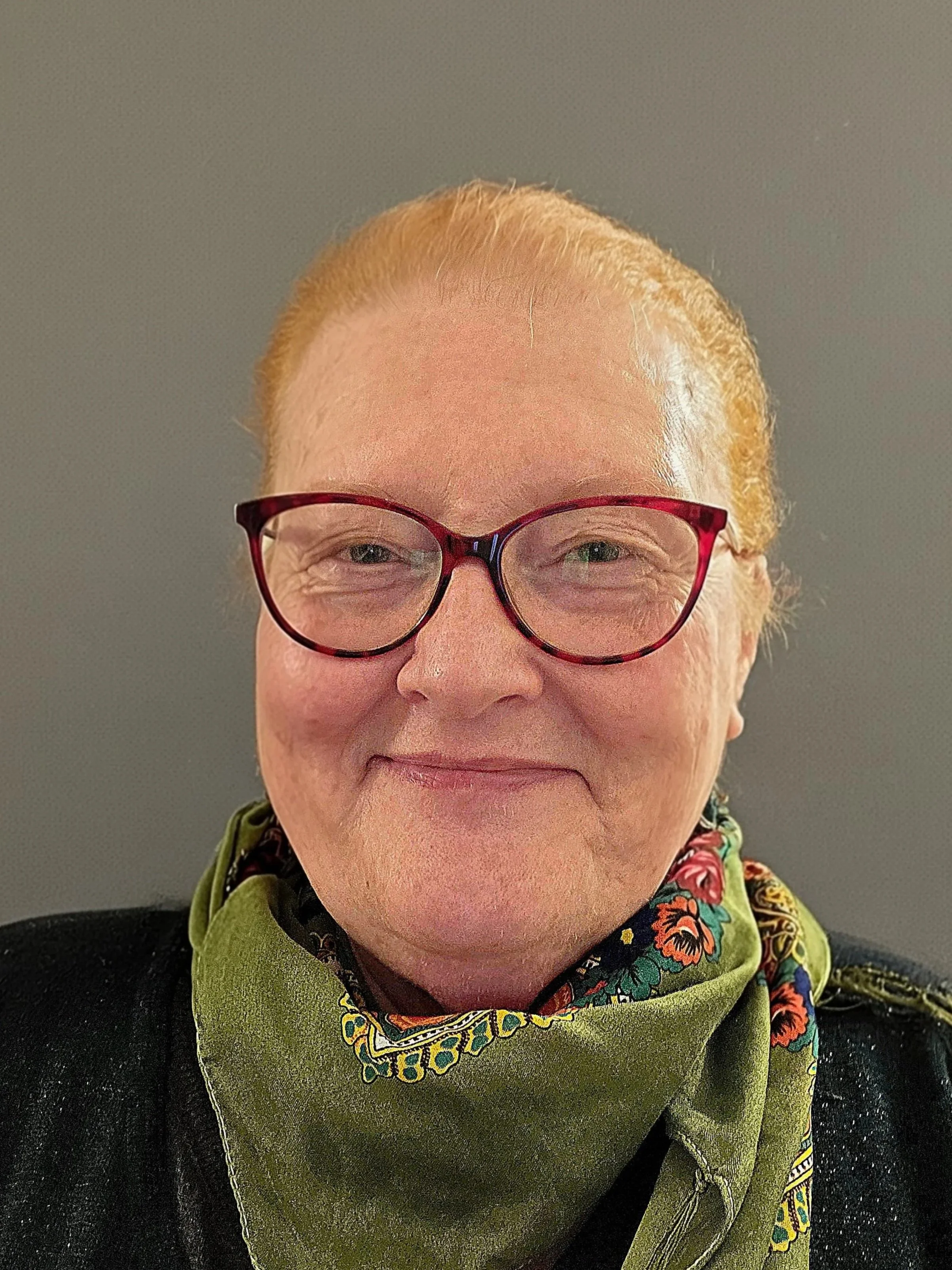 Close-up of a woman with red hair wearing glasses and a green bandana, smiling against a neutral background.