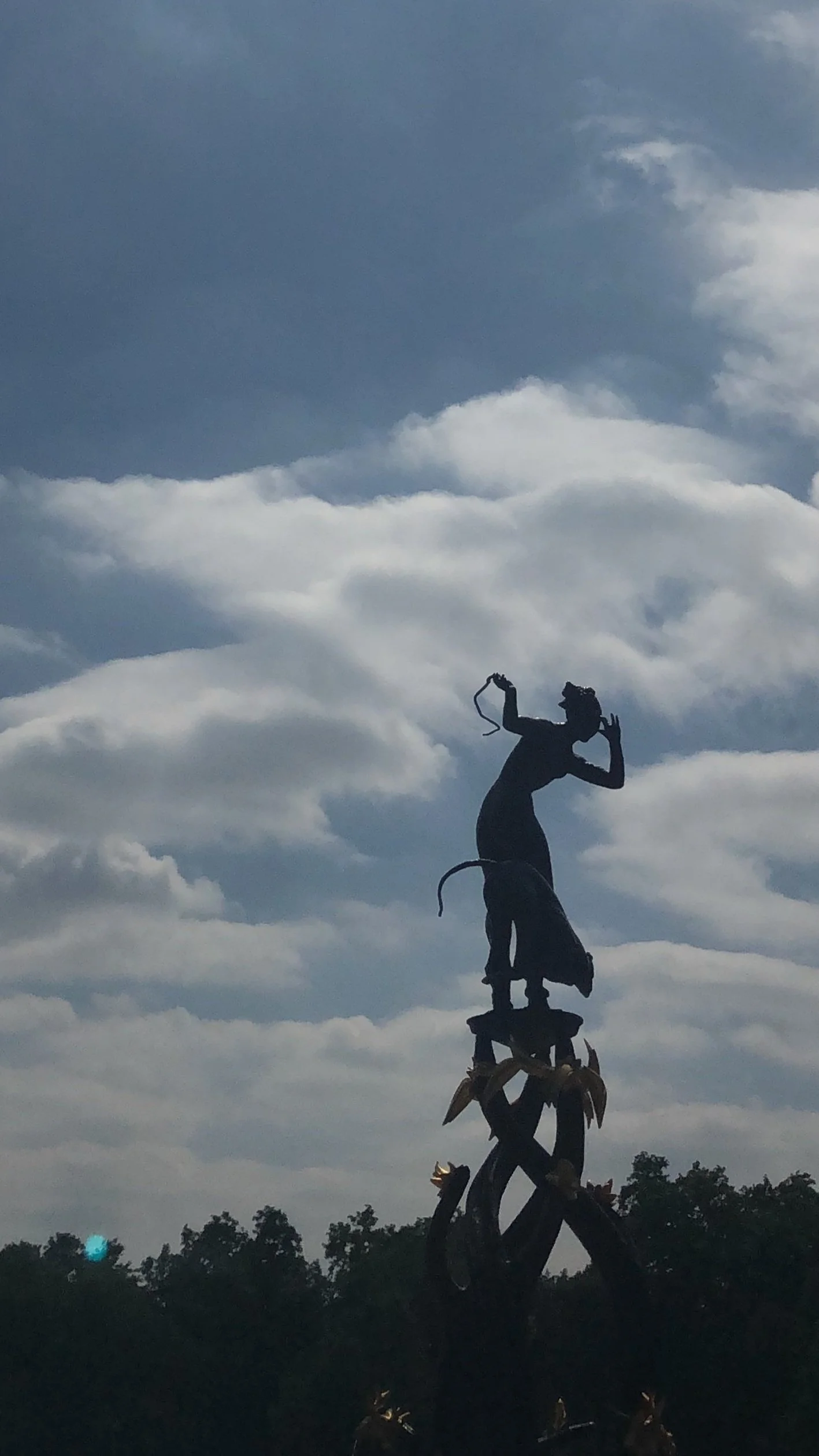 Silhouette of a tall African statue of a woman holding a necklace, standing on a decorated pedestal against a partly cloudy sky.