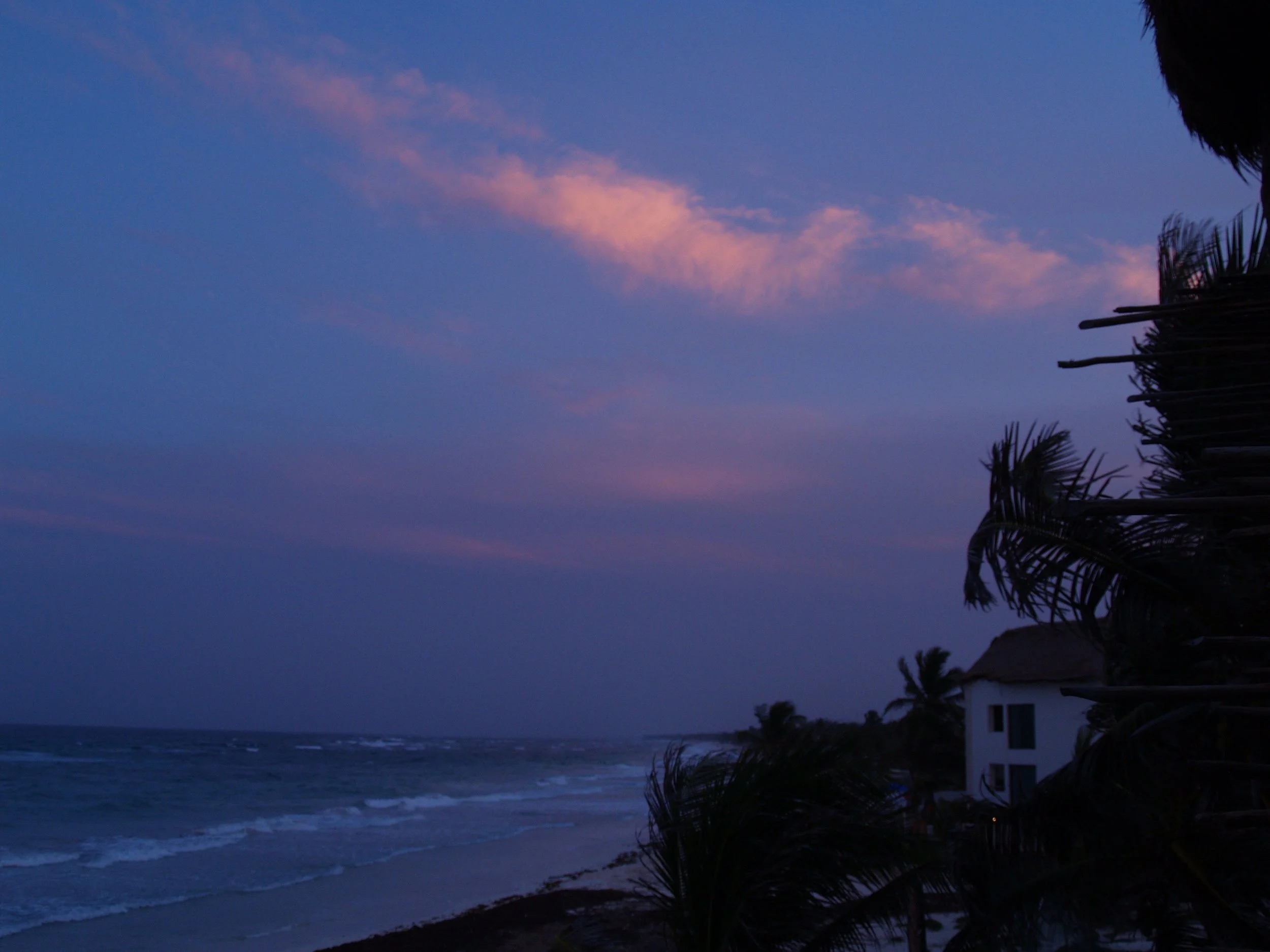 A beachside scene at dusk with purple and pink clouds in the sky, waves hitting the shore, and palm trees near a white building.