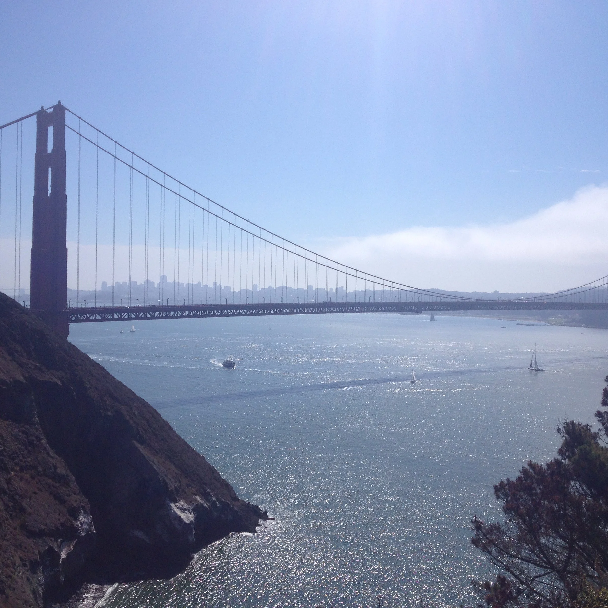 A view of the Golden Gate Bridge over a body of water with sailboats, a boat, and a distant cityscape in the background, under a partly cloudy sky.
