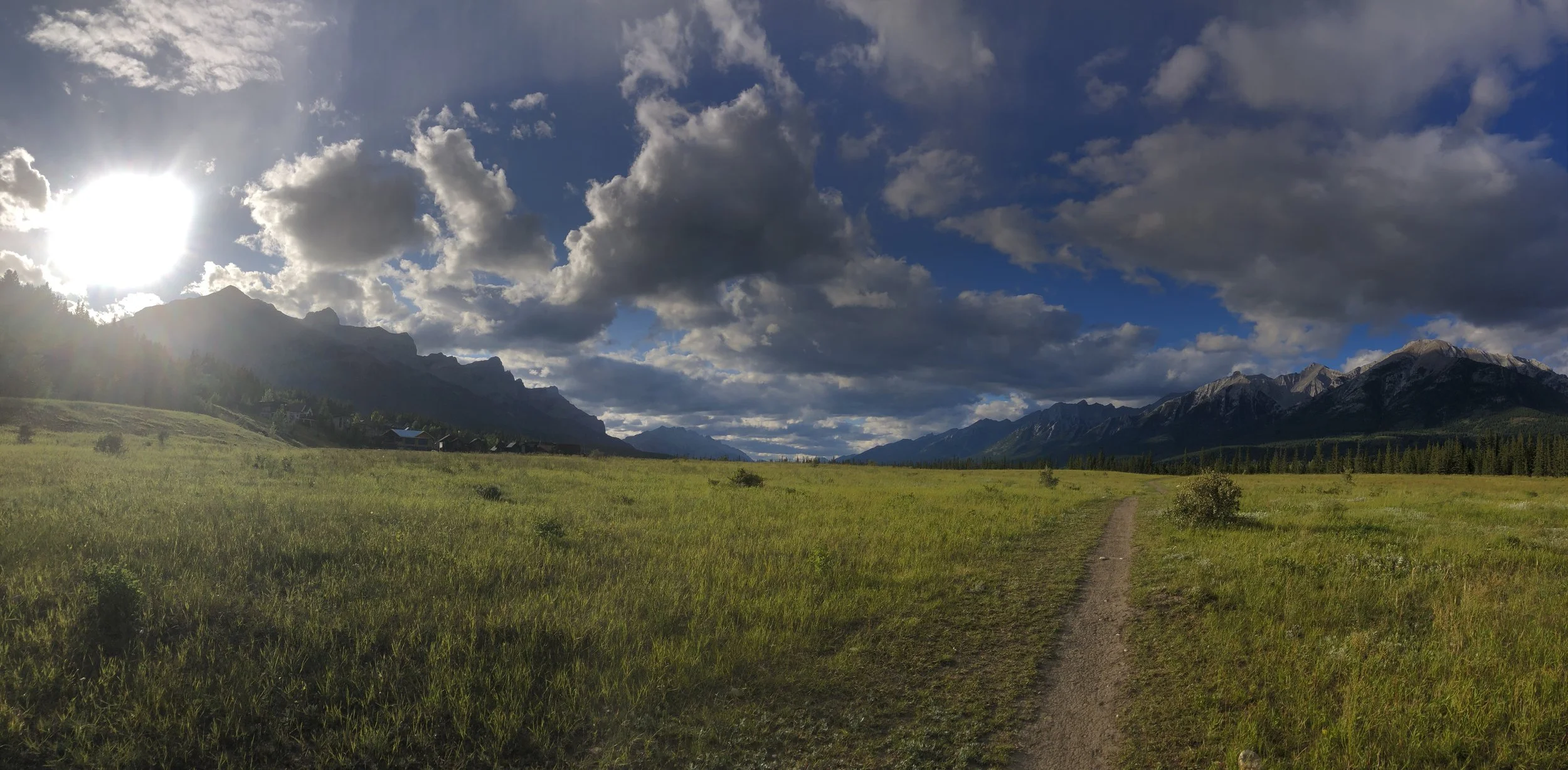 A scenic landscape with a narrow dirt path through a grassy field, mountains in the background, and a partly cloudy sky with the sun peeking through.