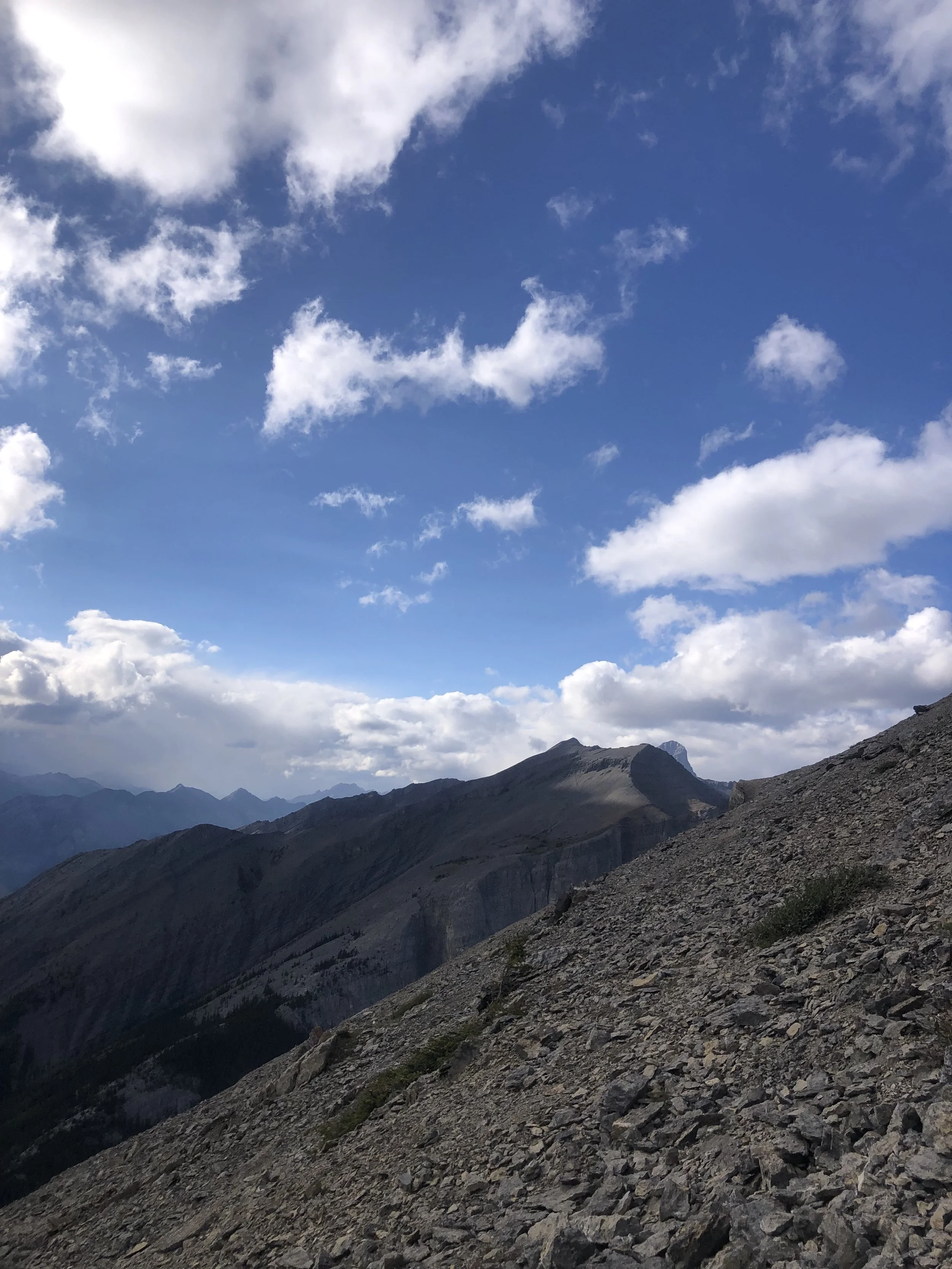 Mountain landscape with rugged terrain in the foreground, multiple mountain peaks in the background, and a partly cloudy blue sky above.