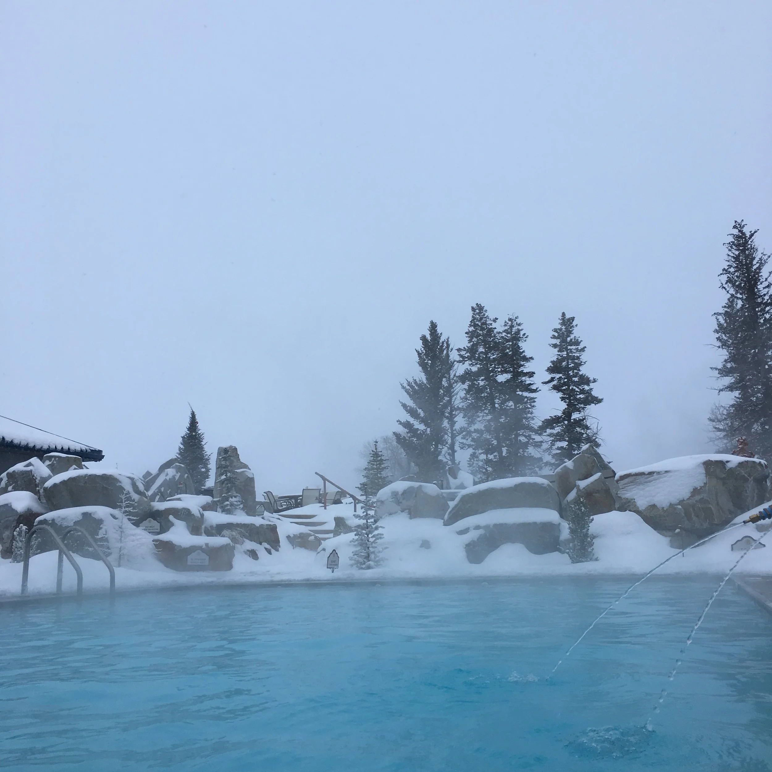 Outdoor hot spring pool surrounded by snow-covered rocks and trees, with misty mountains in the background.