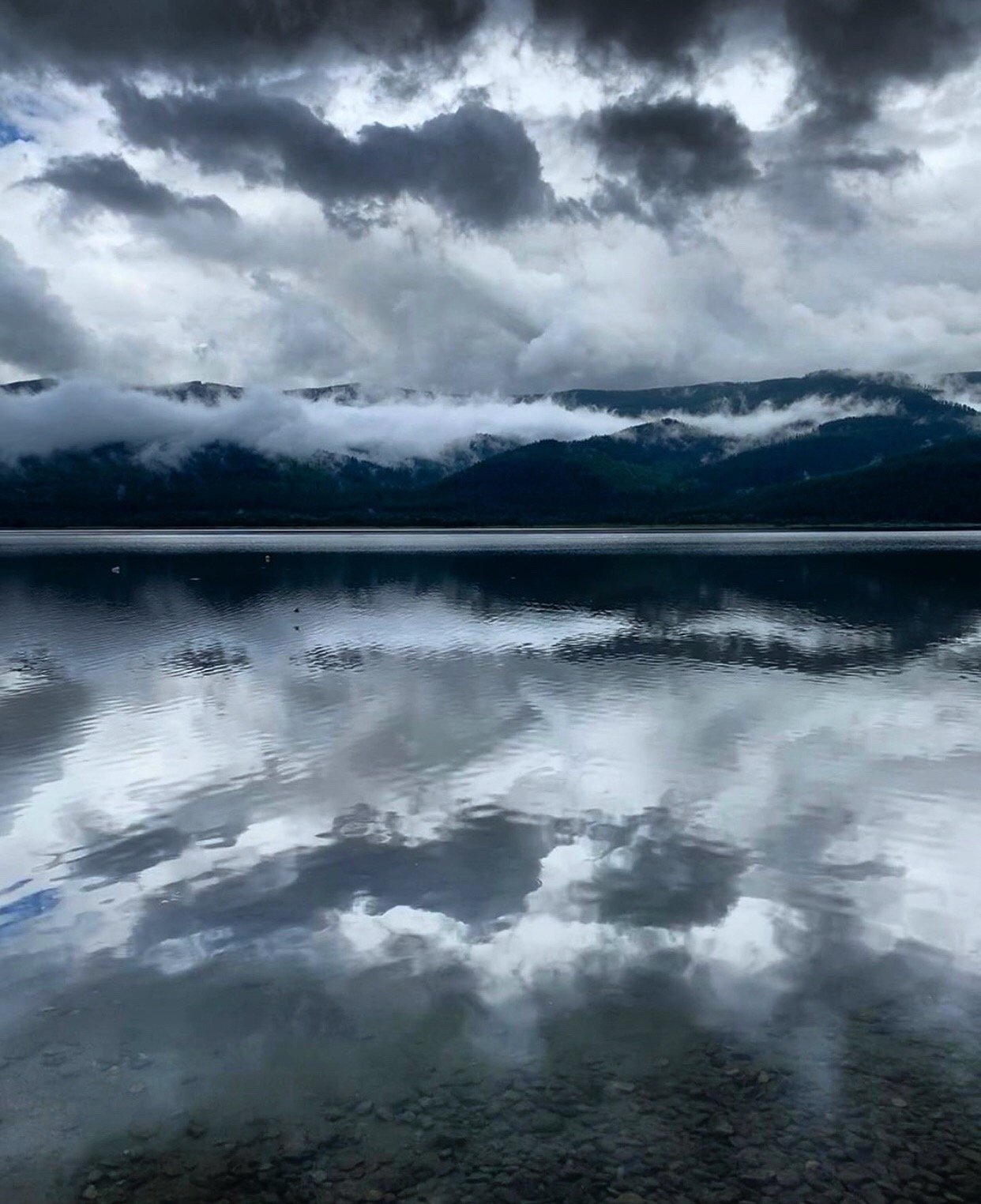 Cloudy sky with mountains partially covered by fog reflected in a calm lake with a rocky bottom.