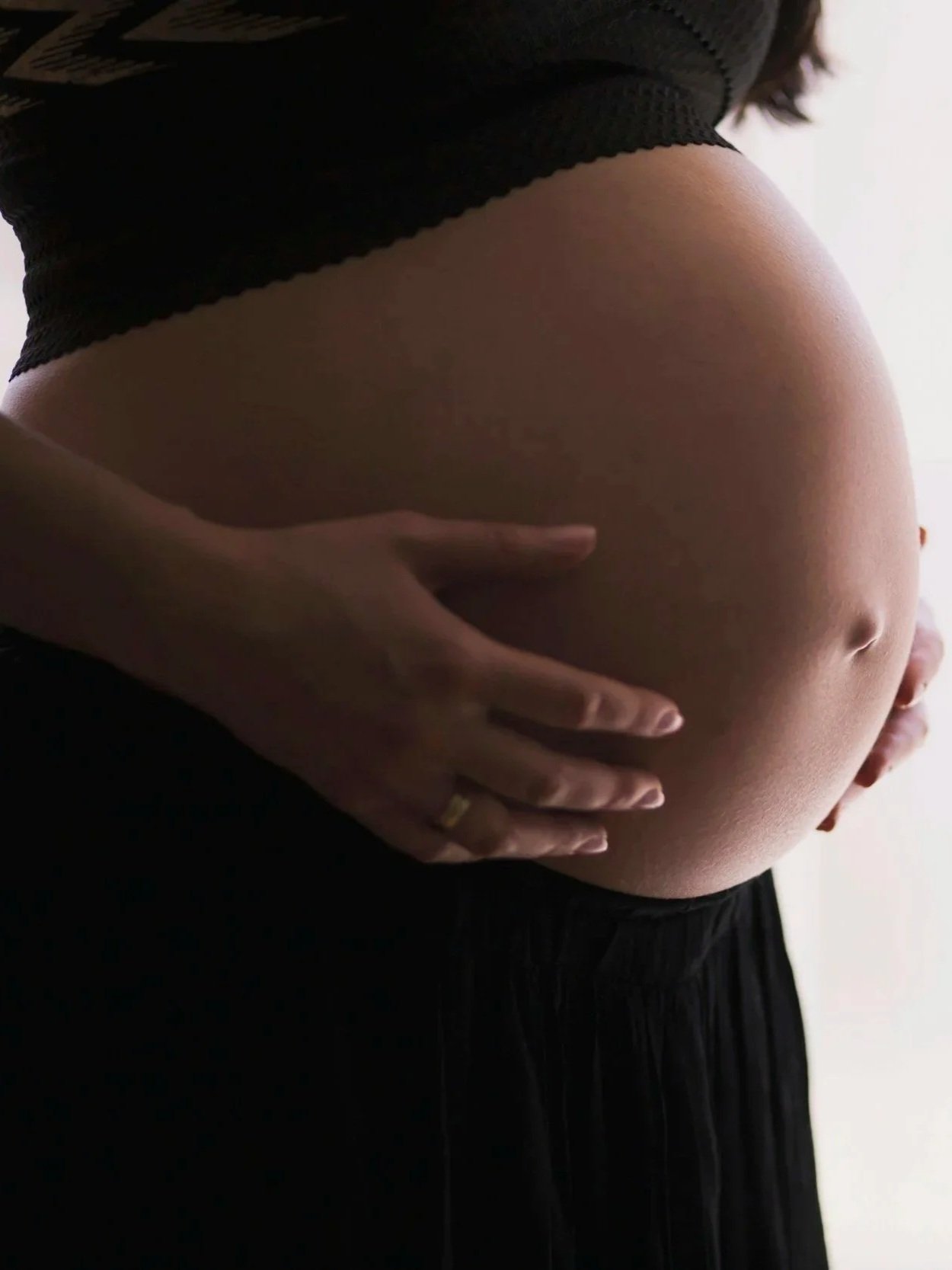 Close-up of a pregnant woman gently cradling her belly with both hands. She is wearing a black top and skirt. The focus is on her pregnant belly and hands.