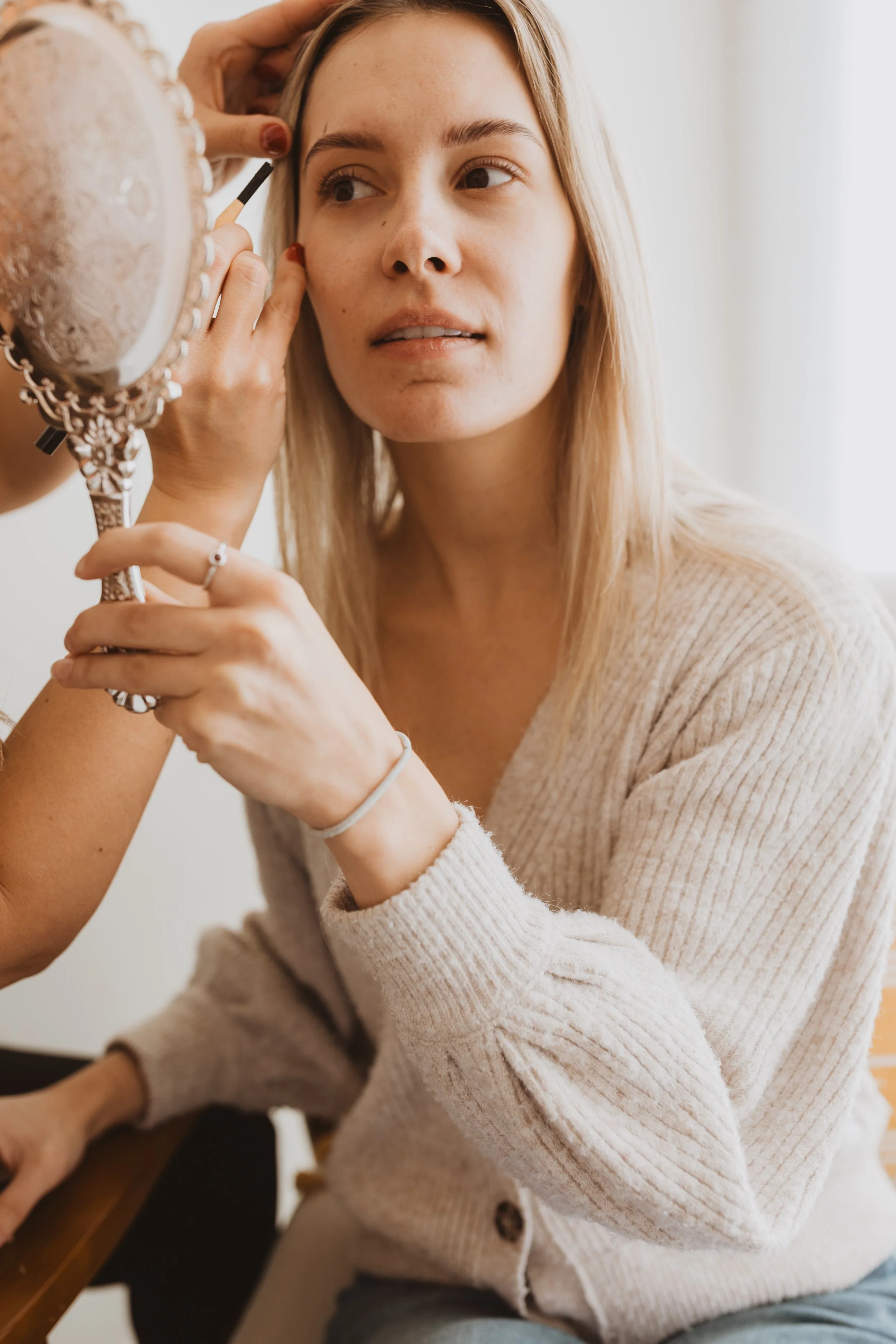 A woman sitting at a table getting her makeup done, looking into a hand mirror with a neutral expression.
