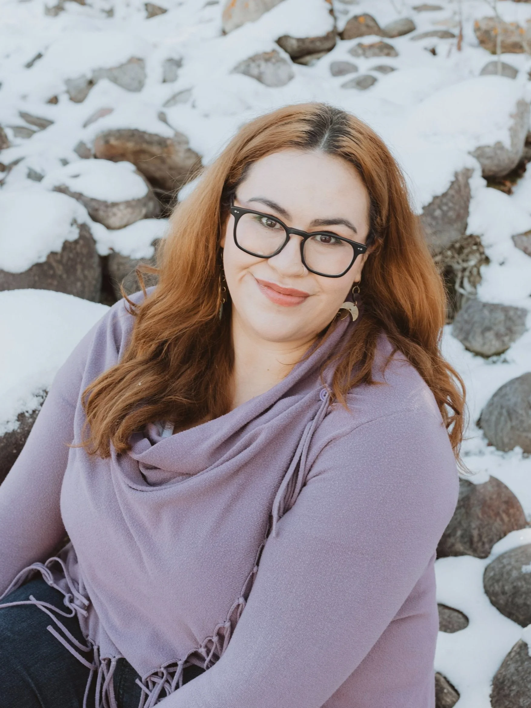 A woman with long, wavy auburn hair and glasses sitting outdoors with snow-covered rocks in the background.