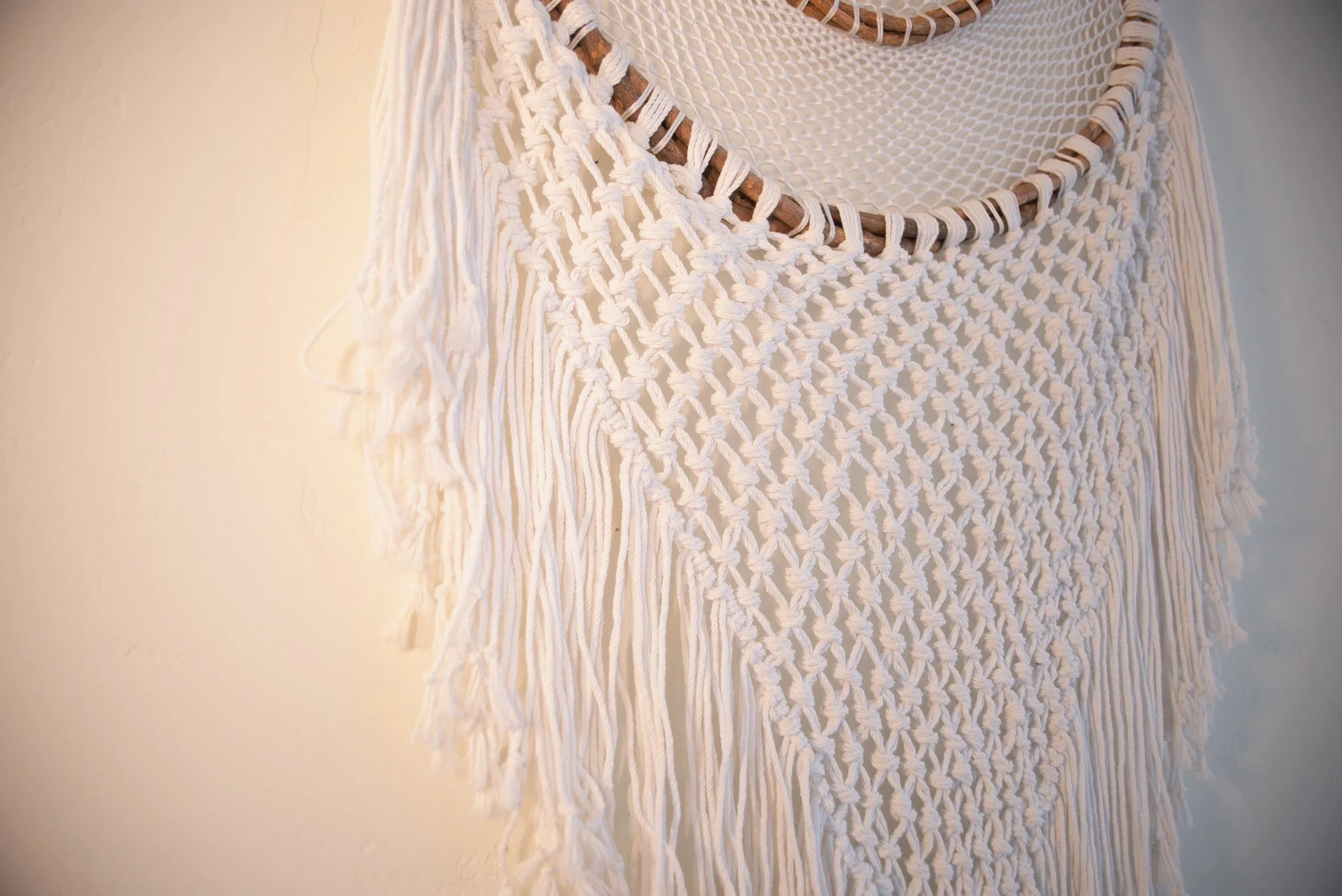 Close-up of a cream-colored macrame wall hanging with fringes, hanging on a light-colored wall.