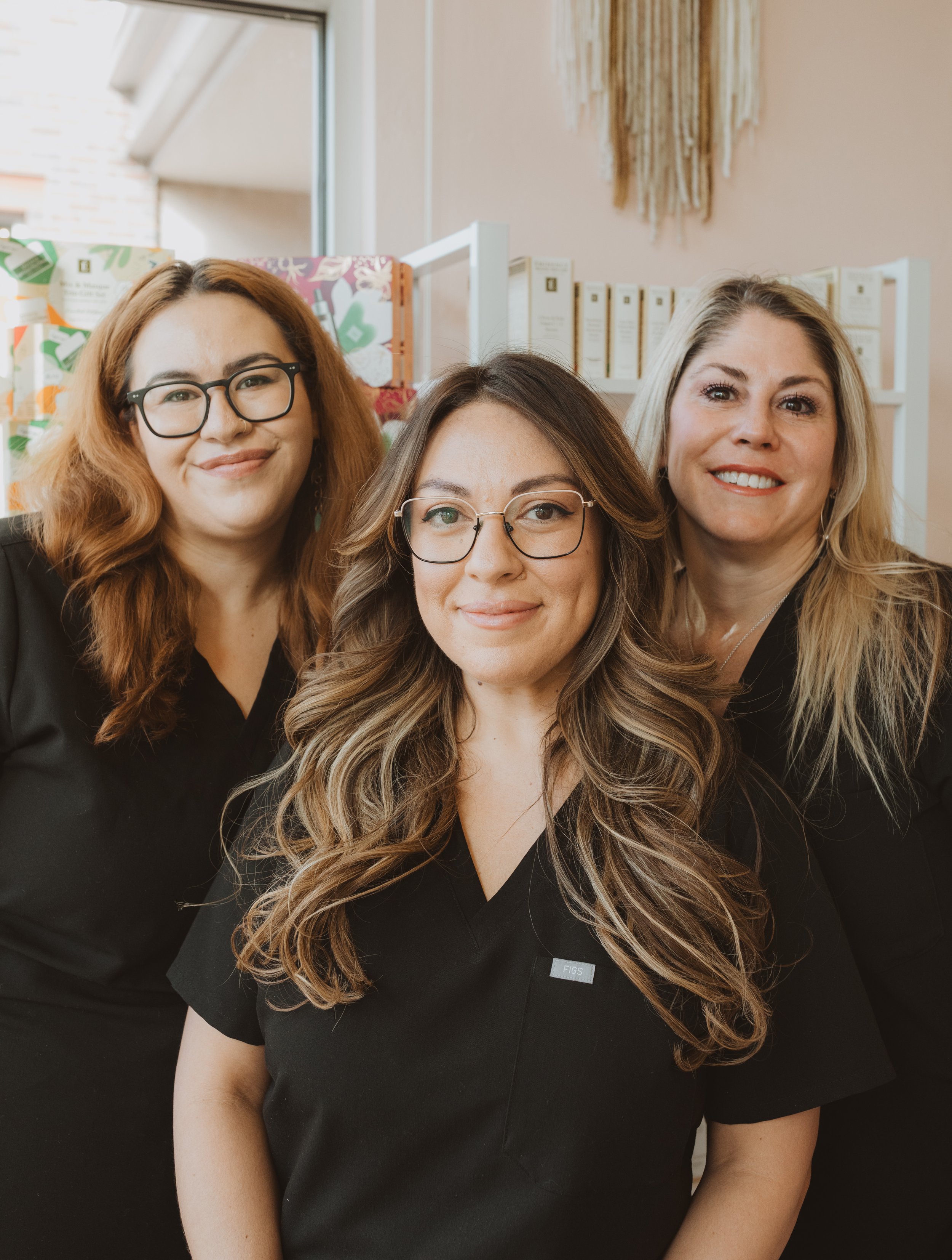 Three women wearing black uniforms standing together indoors with shelves and decorative wall hanging in the background.