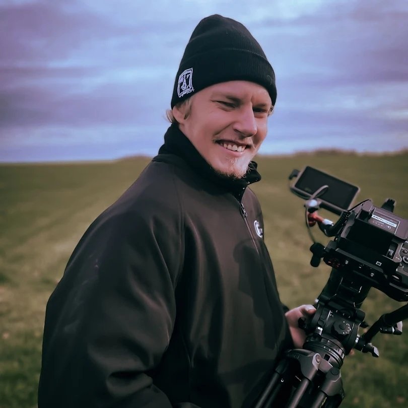 Smiling man in black beanie and jacket holding a professional video camera outdoors on a grassy field with overcast sky.